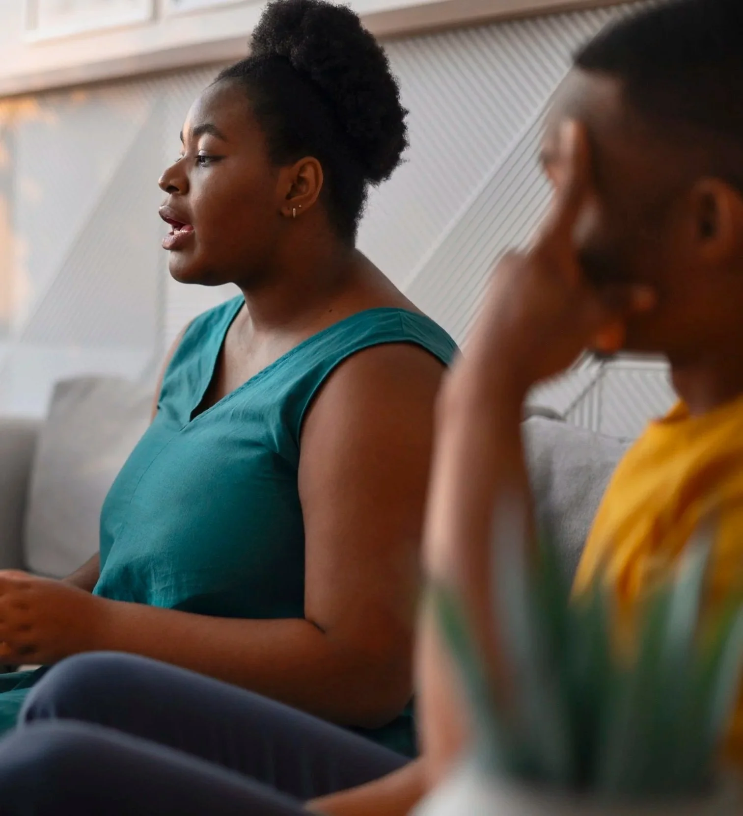 Two women sitting on a couch engaged in conversation, one in a teal sleeveless top with natural hair styled in an updo, the other in a yellow top with short hair, blurred in the foreground.