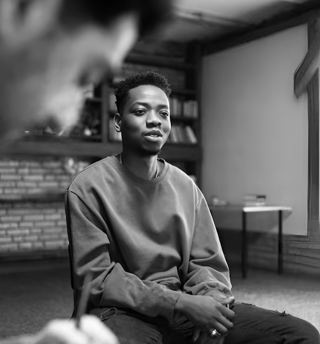 A young man with short hair sitting indoors, appears to be having a conversation, with a blurred person in the foreground, in a room with a brick fireplace and bookshelves.