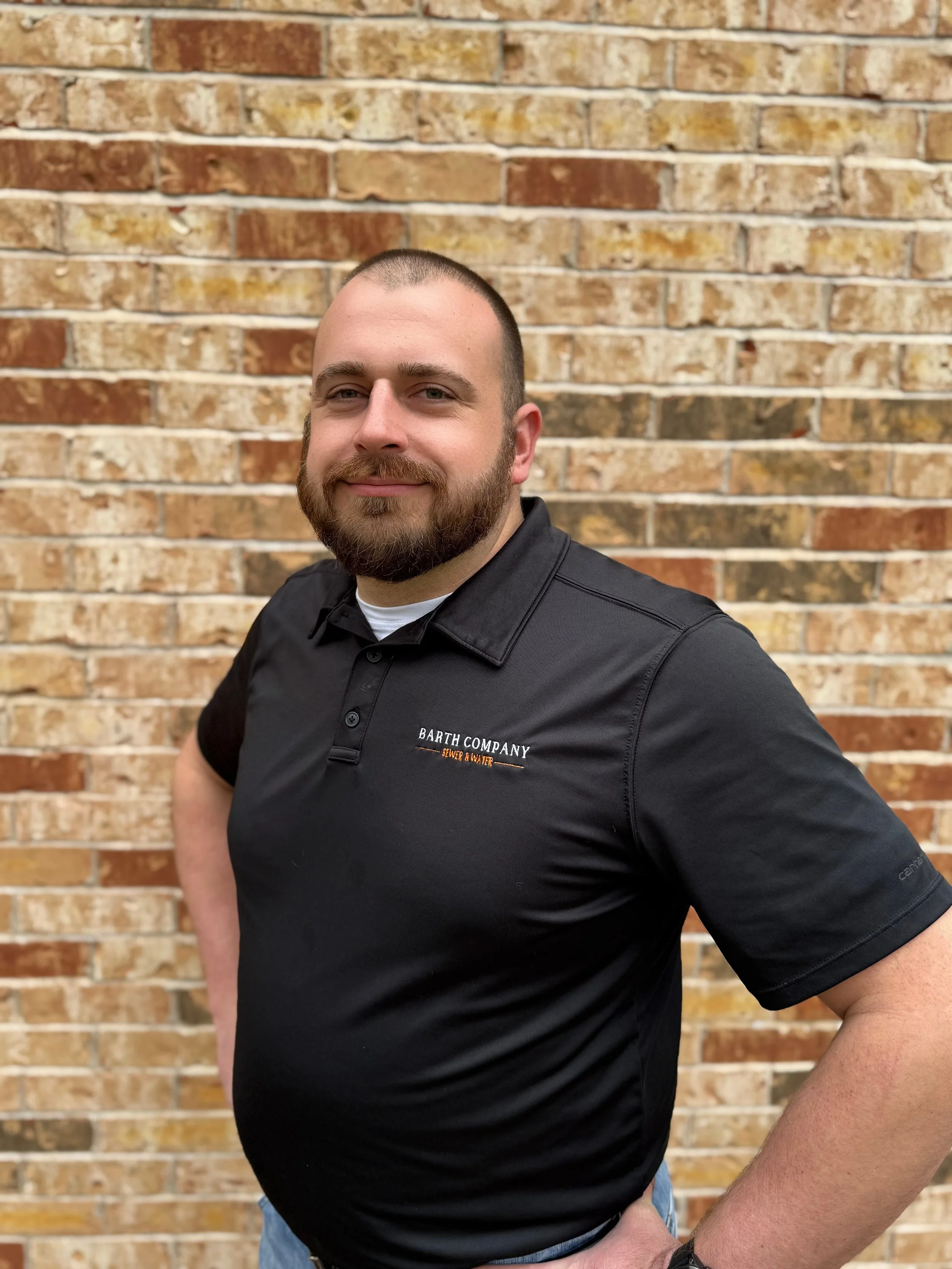 A man with a beard and short hair wearing a black polo shirt standing in front of a brick wall.
