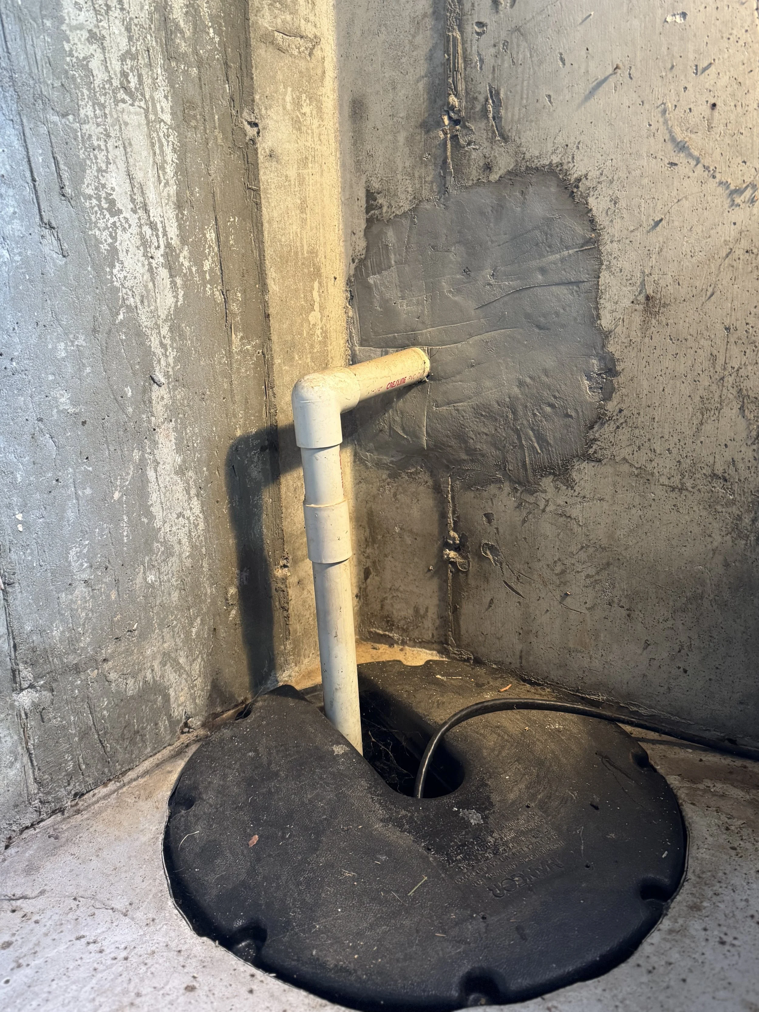 A squat toilet with a black rubber matt surrounding it, installed in a concrete floor with unfinished concrete walls. A white PVC pipe is positioned in the bowl of the toilet.
