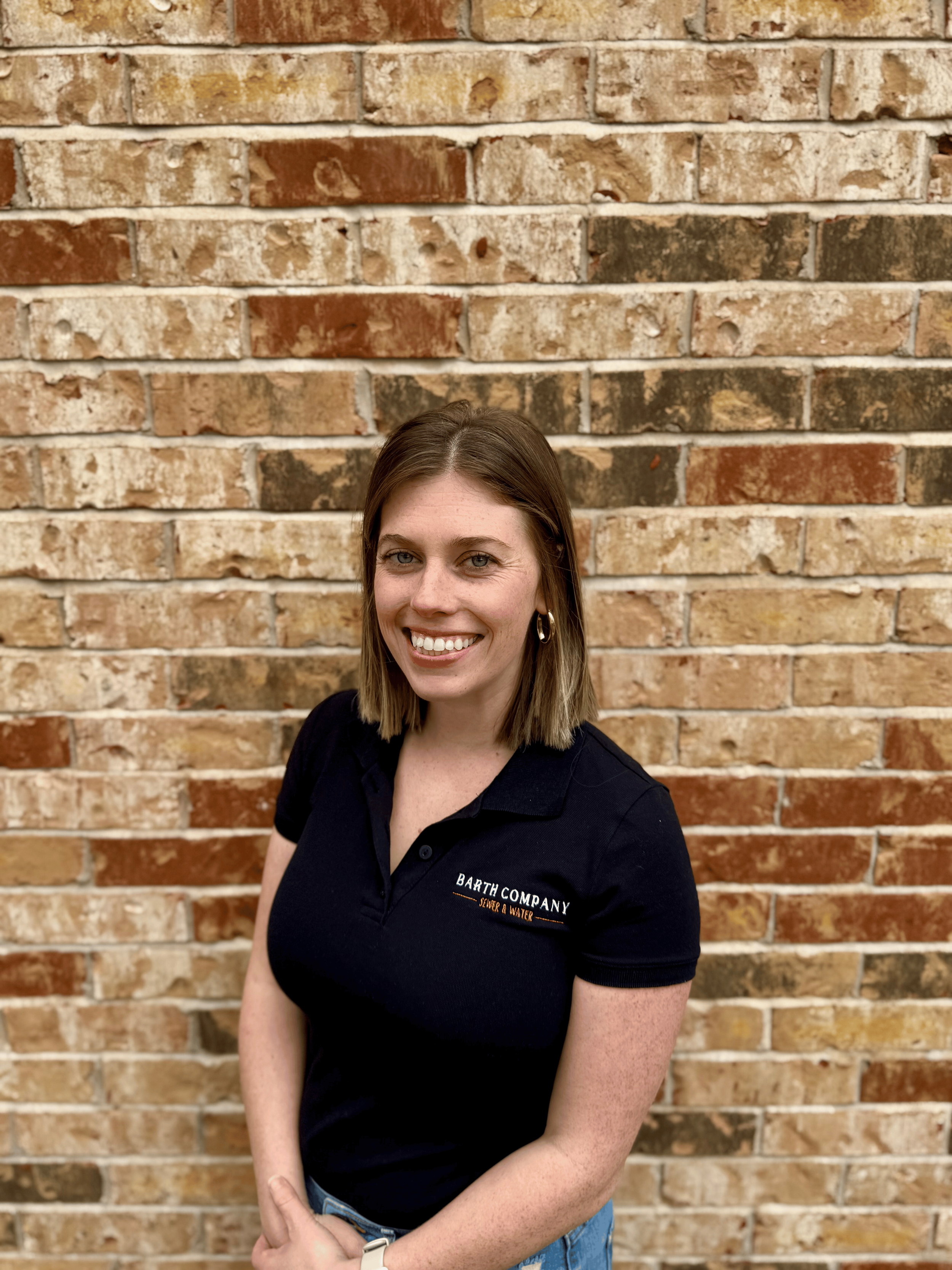 A woman with shoulder-length brown hair smiling, wearing a black polo shirt with "Barth Company" logo, standing in front of a brick wall.