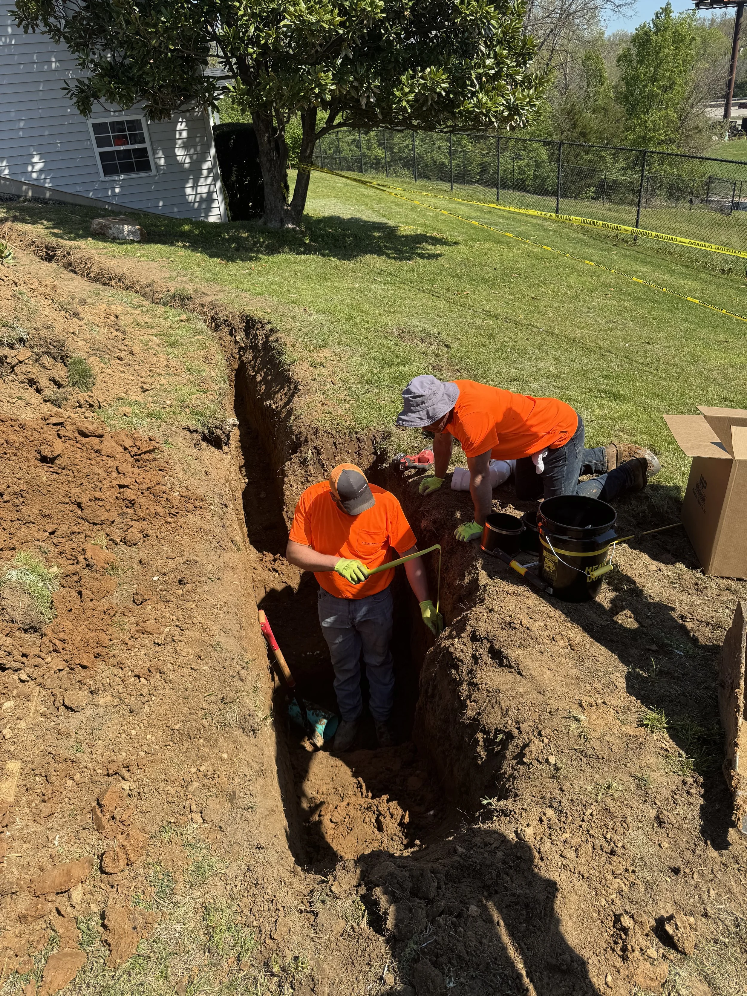 Workers in orange shirts digging a trench in a yard near a house with a white exterior. They are using shovels and working inside the trench, which is surrounded by disturbed earth. A yellow caution tape and a black fence are visible in the background, along with trees and a hint of a road or highway.