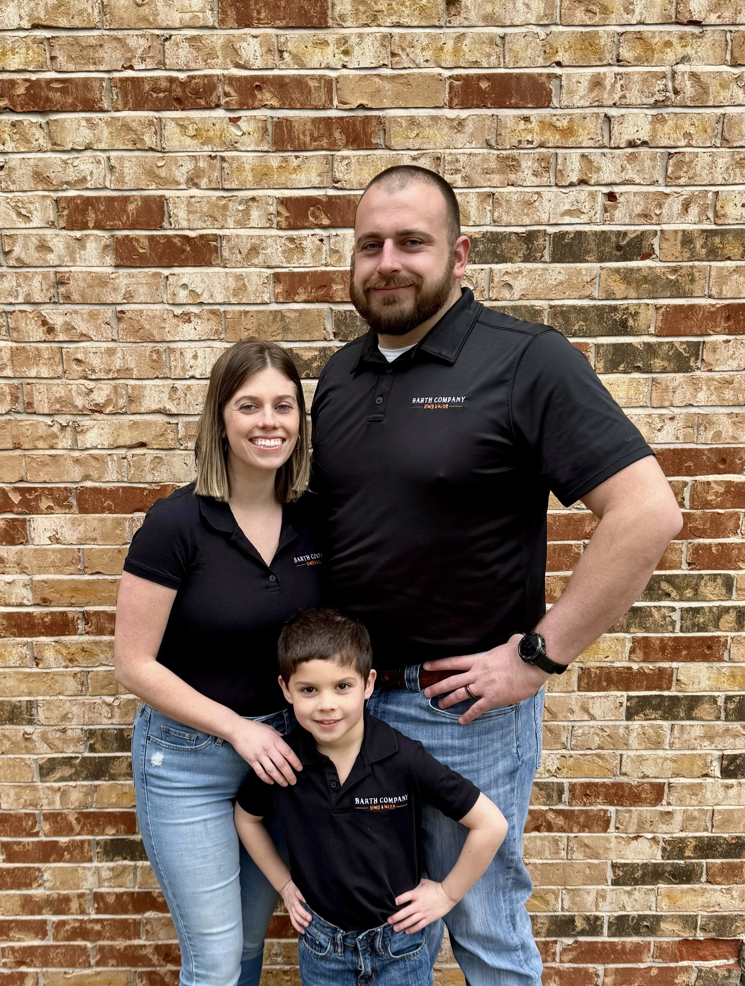A family of three standing in front of a brick wall, wearing matching black shirts with the 'Barth Company' logo.
