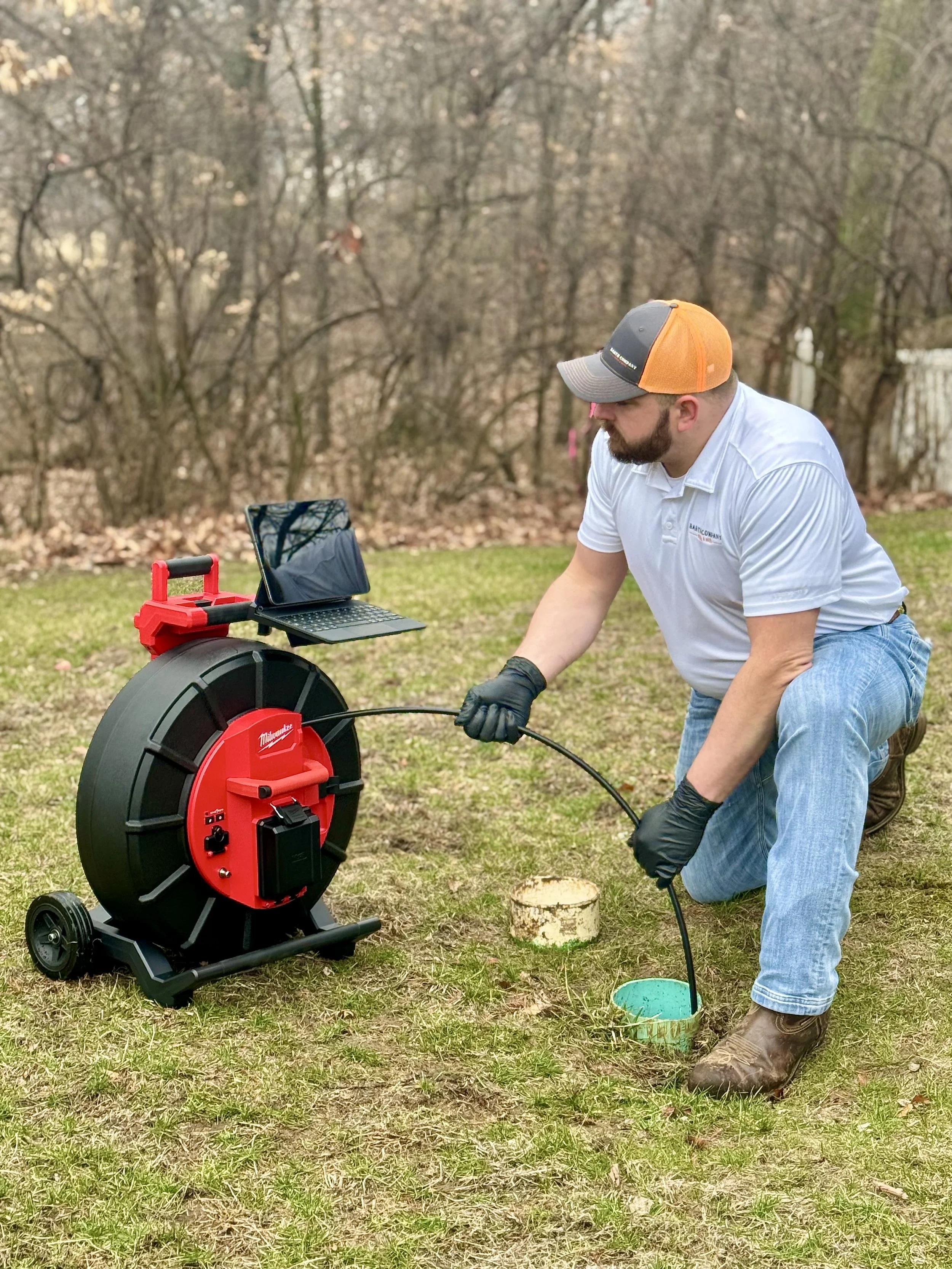 A man kneeling on the grass outdoors next to a sewer inspection camera setup, holding the cable with a tablet placed on a stand nearby, and a tree stump visible in the background.