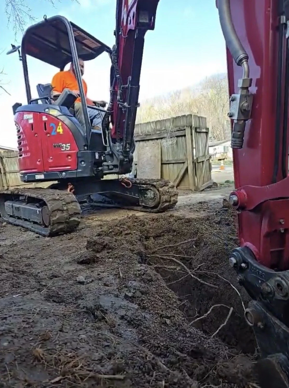 Construction worker operating a red mini excavator digging a trench, with a wooden fence and trees in the background.