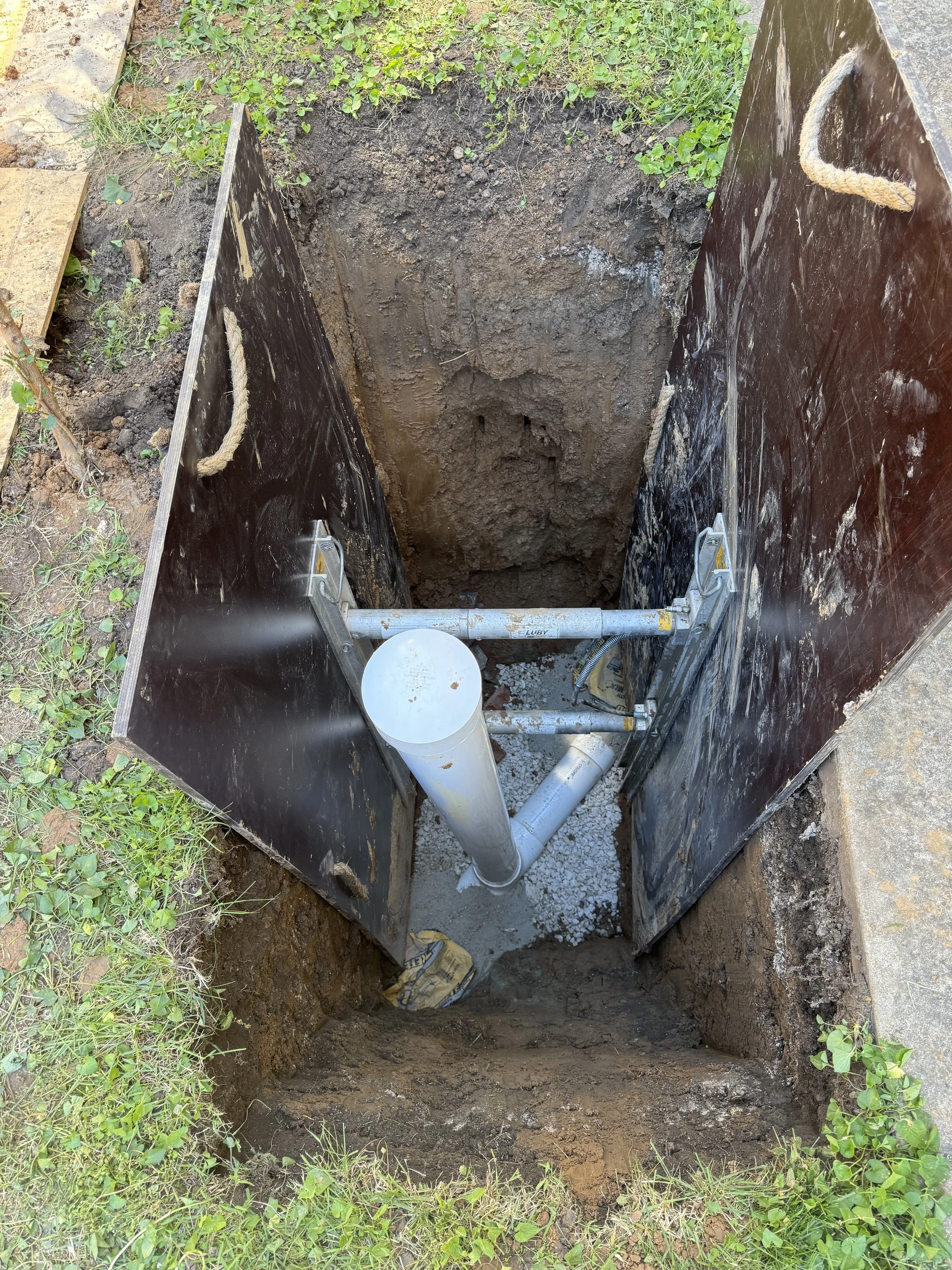An open underground utility access point showing a metal frame, PVC pipes, and electrical wiring inside a dirt excavation site, surrounded by grass and soil.