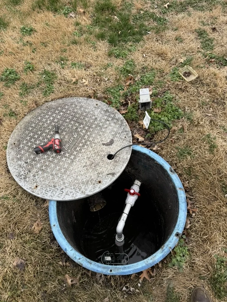 Access hatch open to reveal a water tank with a PVC pipe connected inside, on grassy ground with a cordless drill on the hatch cover and electrical wiring nearby.