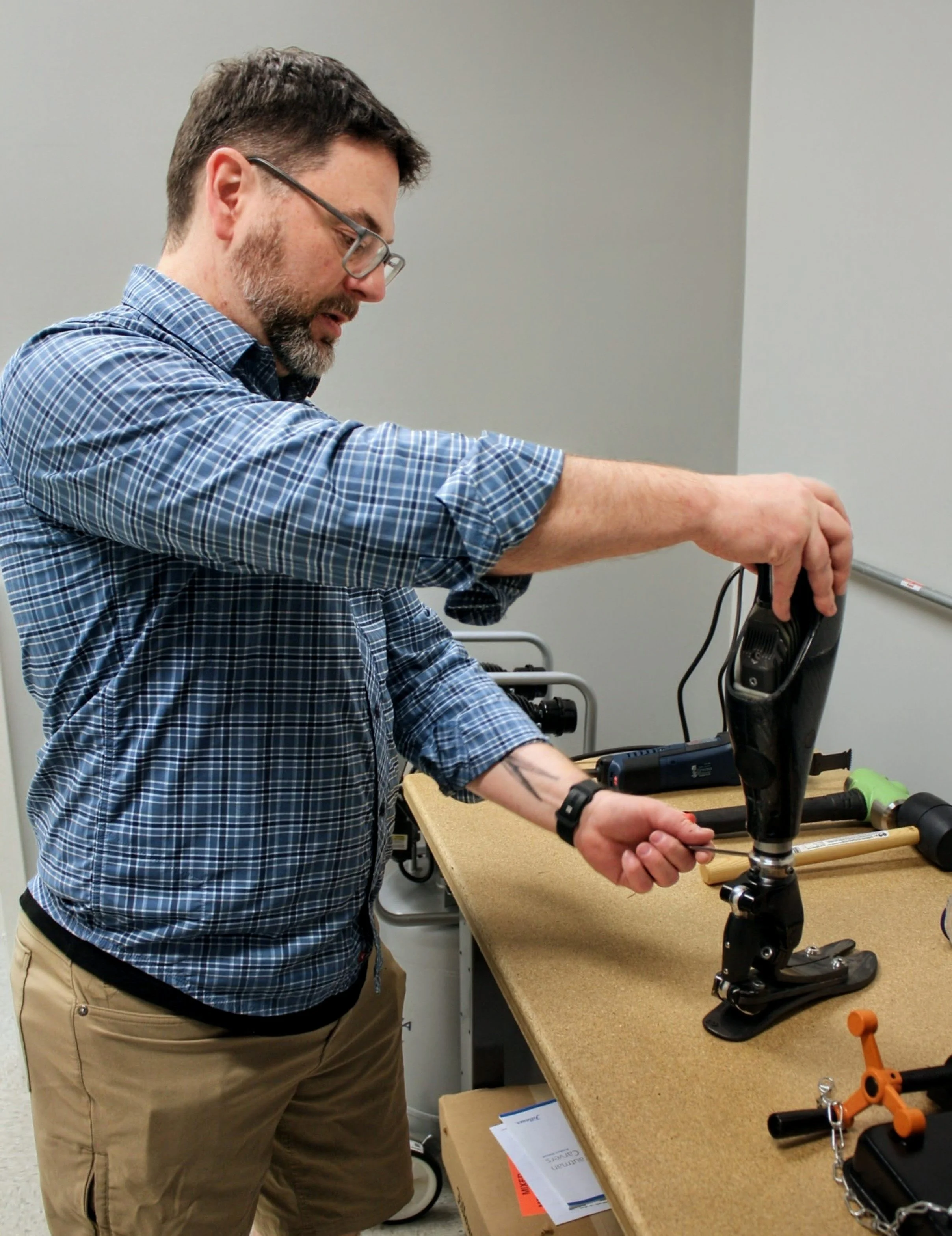 A man in a plaid blue shirt and khaki shorts is working on a prosthetic leg Otto Bock X3 or similar device on a workbench in a workshop or laboratory.