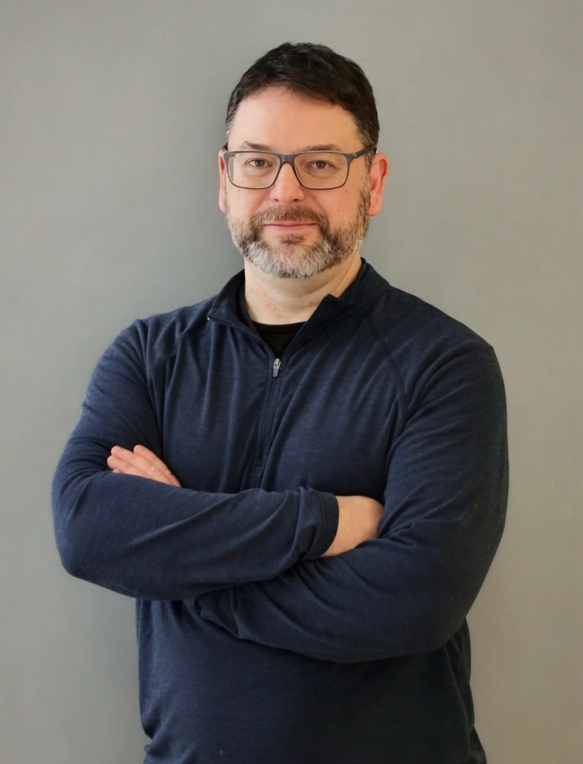 A man with glasses, a beard, and dark hair standing with arms crossed against a plain gray background.