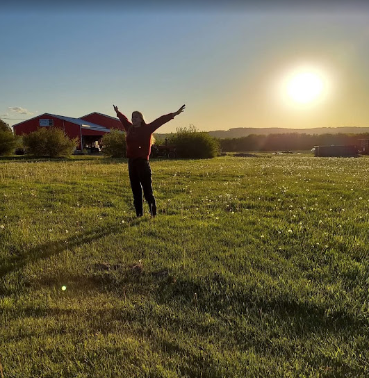 Person standing on grassy field with arms raised at sunset, with buildings and trees in the background.