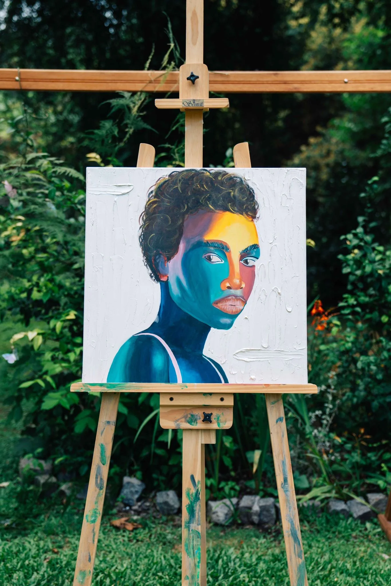 A painting of a woman with curly hair, featuring vibrant blues, oranges, and yellows on her face and shoulders, displayed on an easel against a brick wall.