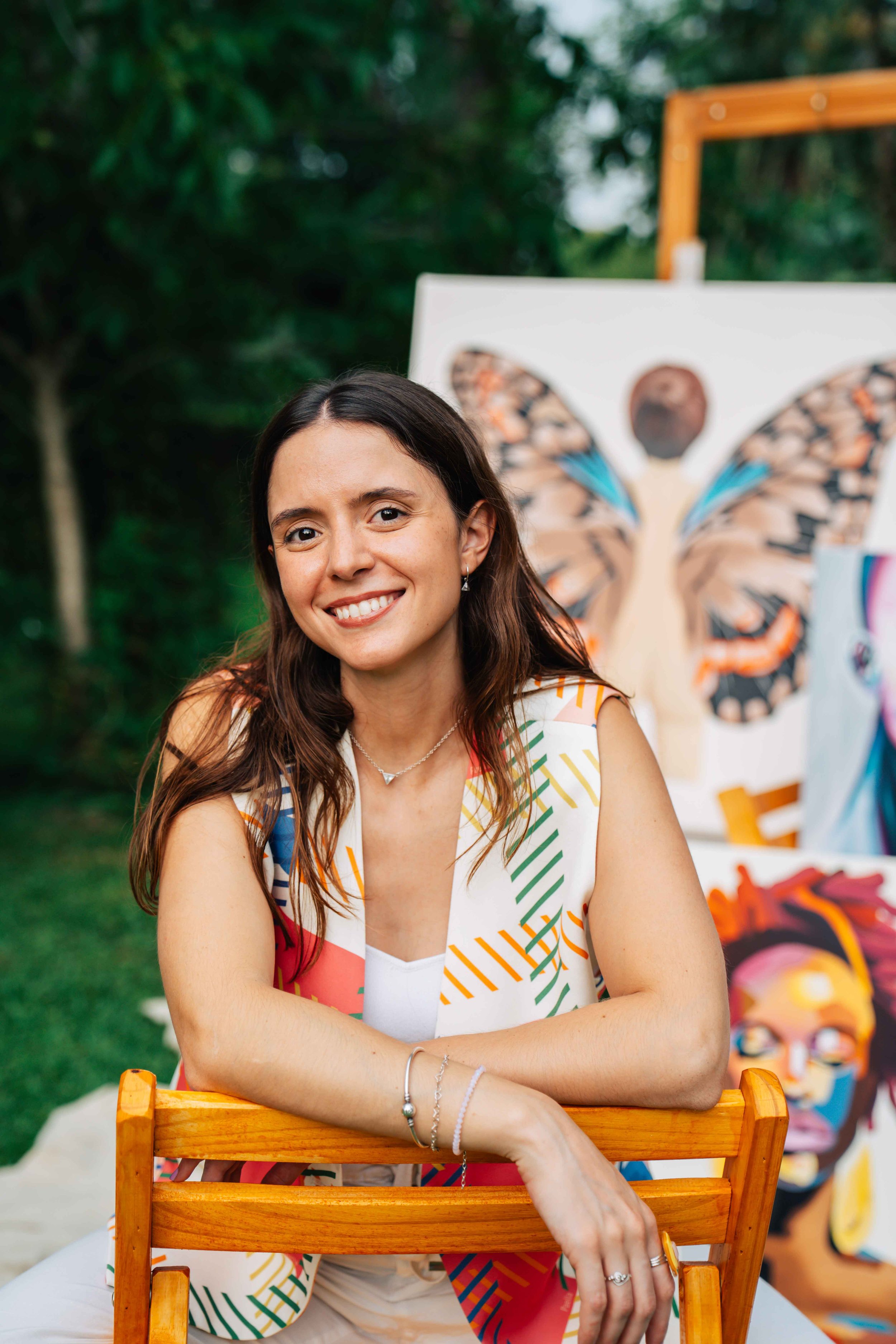A woman with long brown hair and a colorful patterned dress sitting behind a wooden chair outdoors, smiling, with artwork of a butterfly and abstract faces in the background.