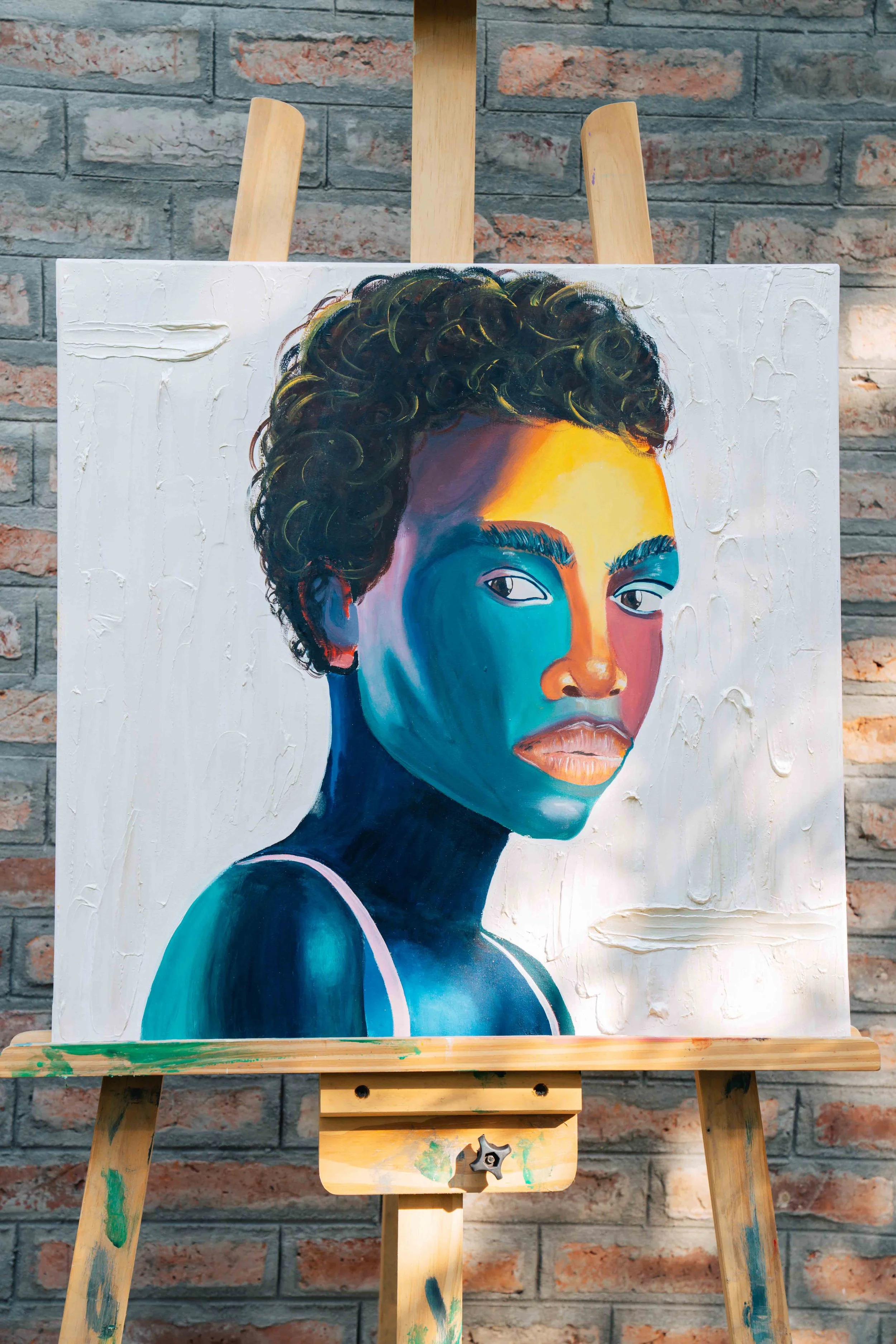 A painting of a woman with curly hair, featuring vibrant blues, oranges, and yellows on her face and shoulders, displayed on an easel against a brick wall.