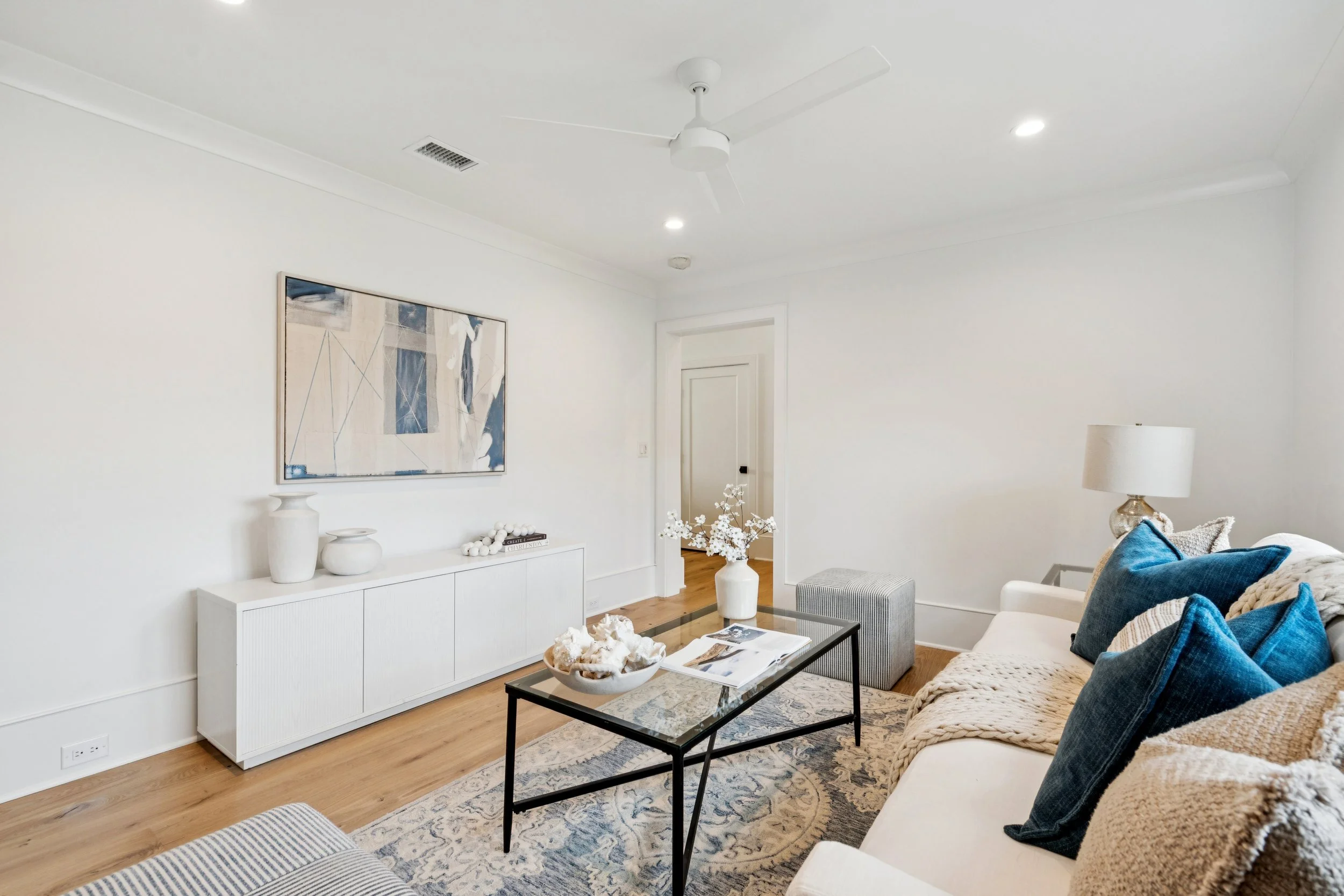 Living room with white walls, hardwood floor, a white sofa with blue and beige pillows, a glass-top coffee table, a white sideboard, a table lamp, and a large abstract painting on the wall.
