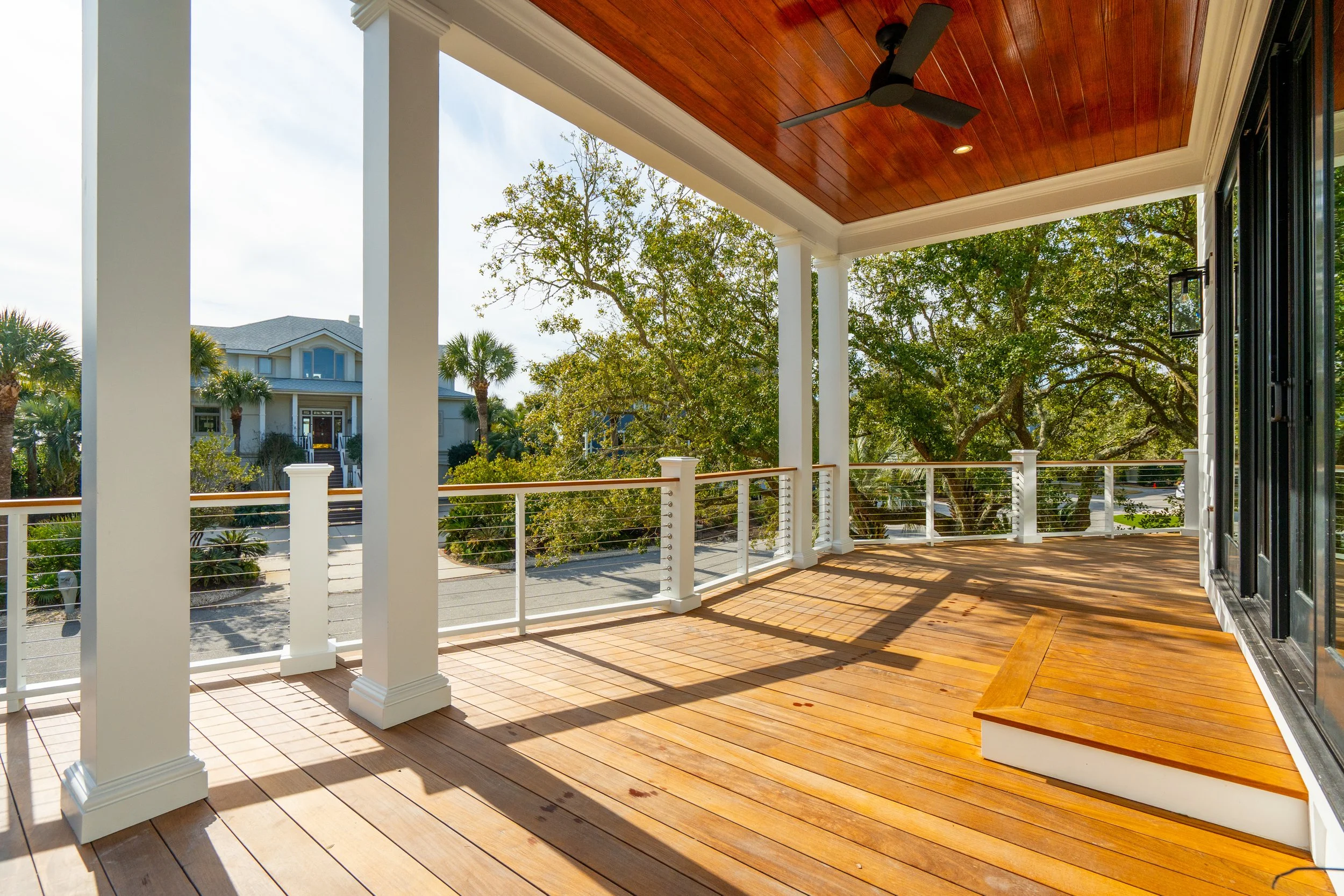 Empty wooden balcony with white railings, ceiling fan, and large sliding glass doors, overlooking a suburban street lined with trees and houses.