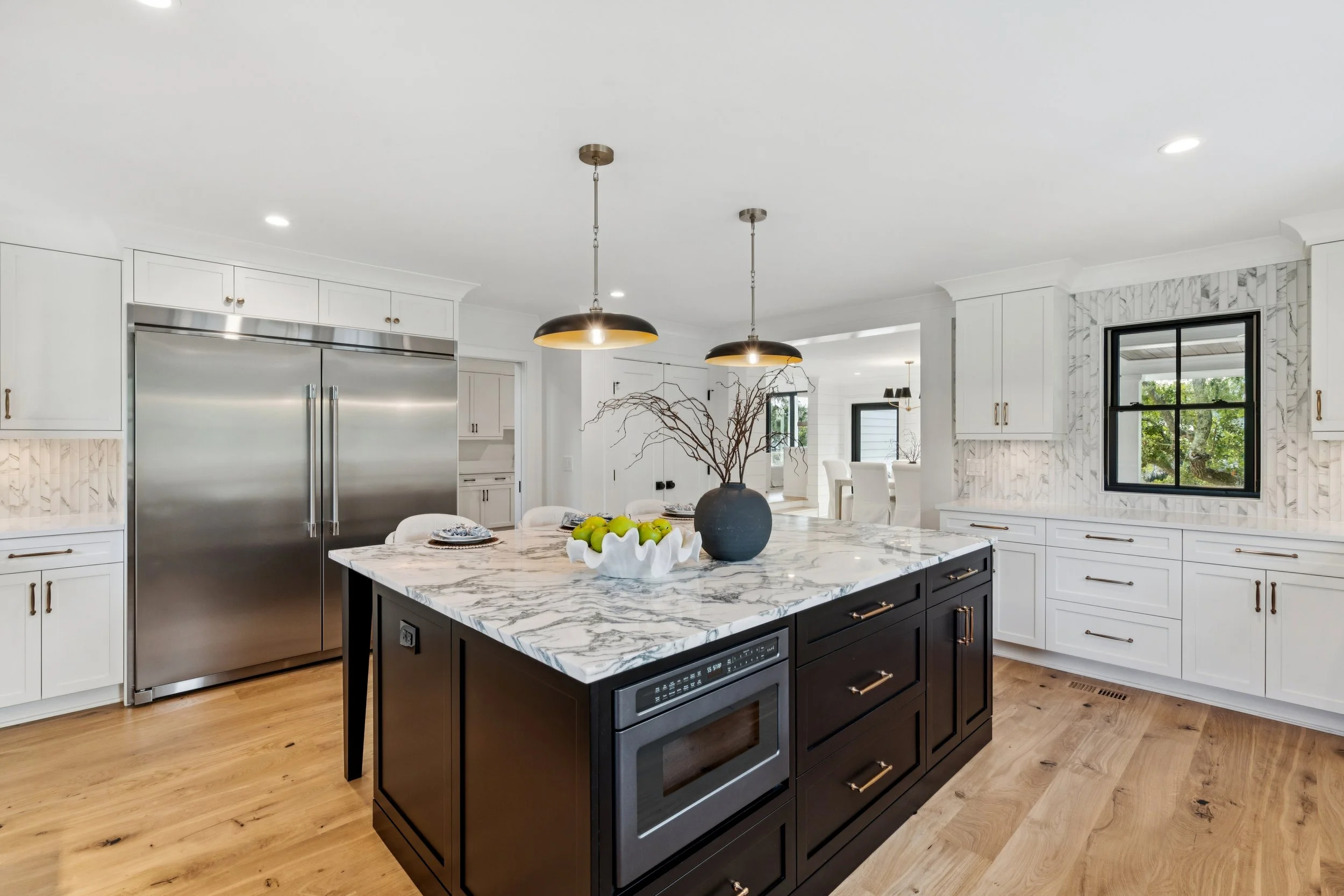 Modern kitchen with a black and white marble island, white cabinetry, stainless steel refrigerator, and a window with greenery outside.