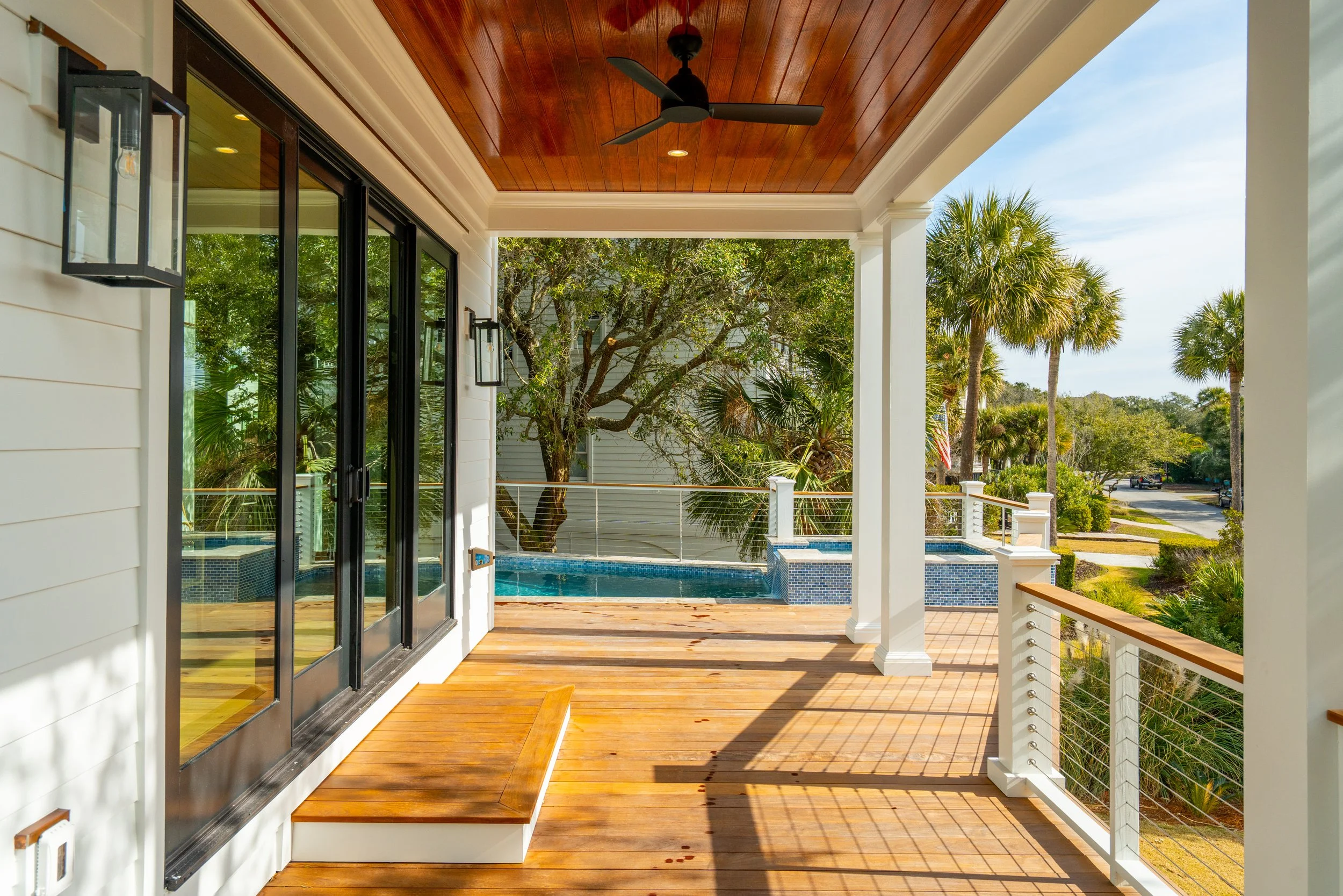 View of a covered porch with wooden flooring, overhead ceiling fan, glass sliding doors, and outdoor lighting fixtures. In the background, there is a small pool or hot tub surrounded by tropical trees and plants, with a neighborhood street visible be