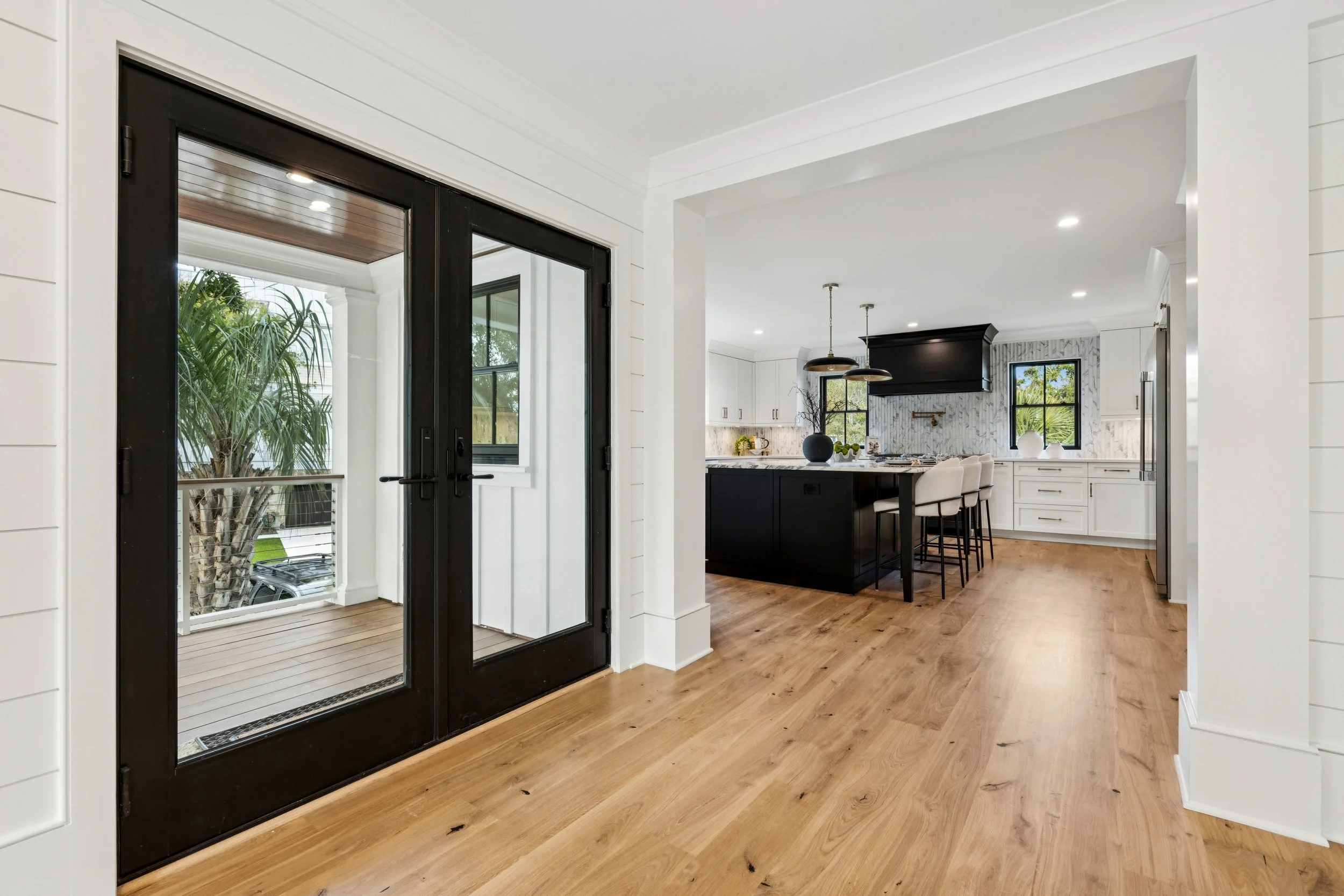 Modern kitchen with black and white cabinetry, a black island, pendant lighting, and hardwood flooring, leading to a covered outdoor patio with glass doors and a view of a green tree.
