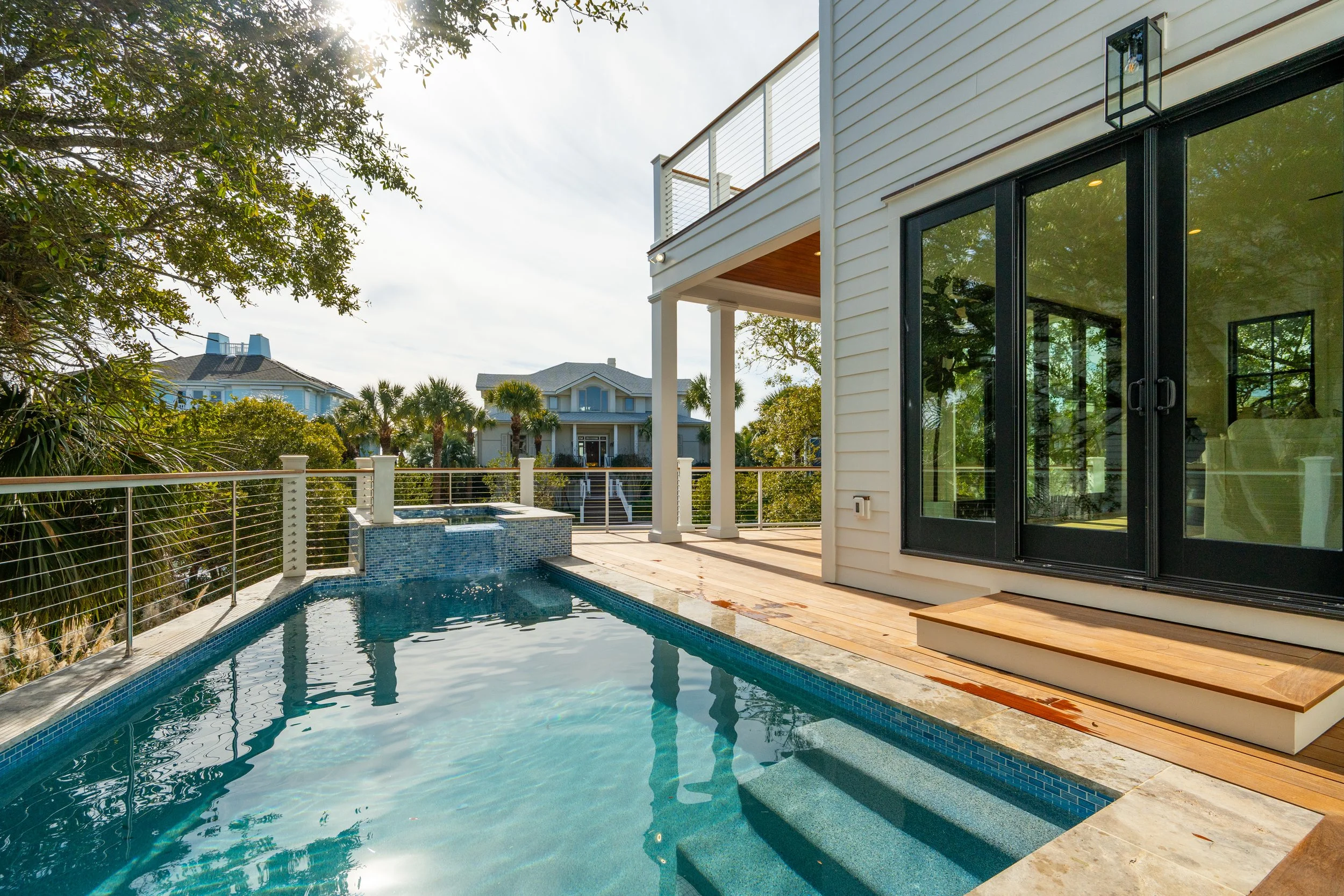 Backyard patio with a small swimming pool, adjacent to a house with sliding glass doors and a deck, with neighboring houses and green trees in the background.