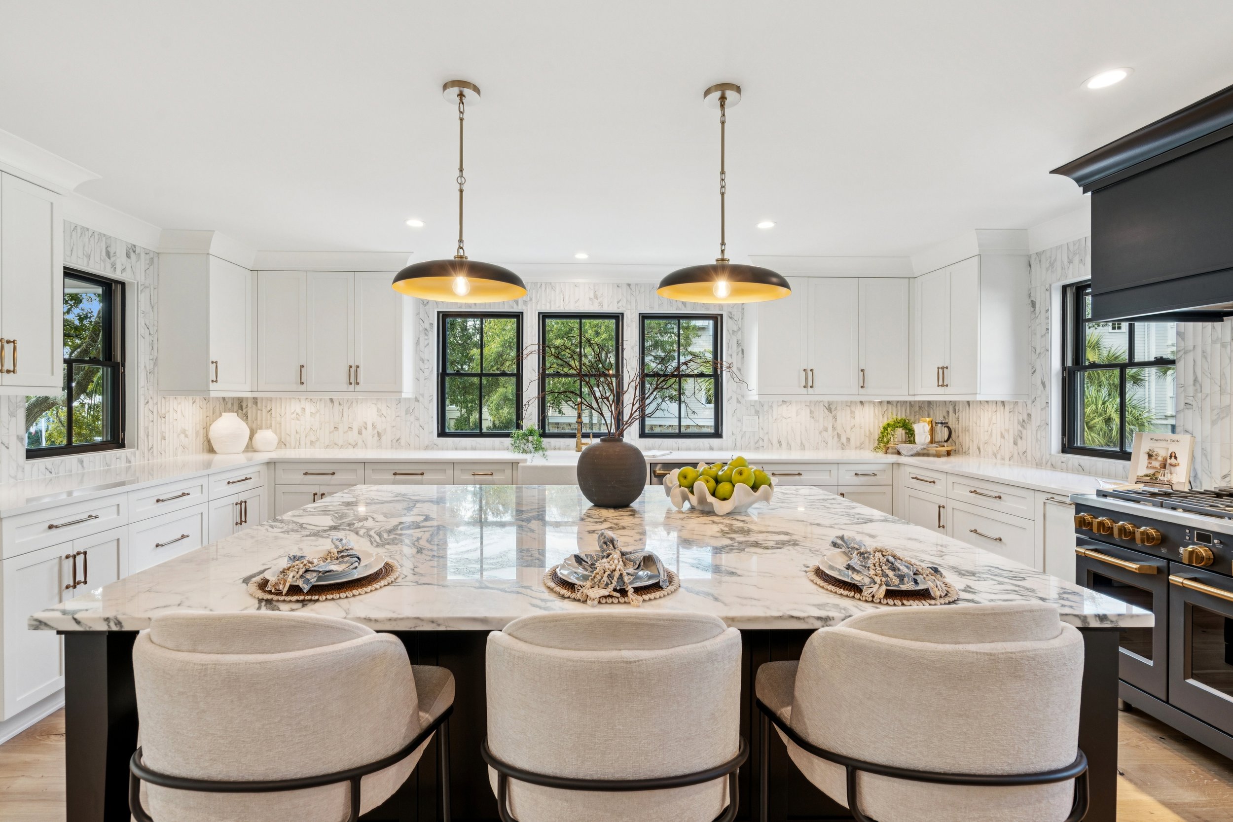 Modern kitchen with white cabinets, marble countertops, and black window frames. An island with a marble top has four beige chairs. Two black pendant lights hang above the island. Decor includes a dark vase with branches, a bowl of green apples, and 