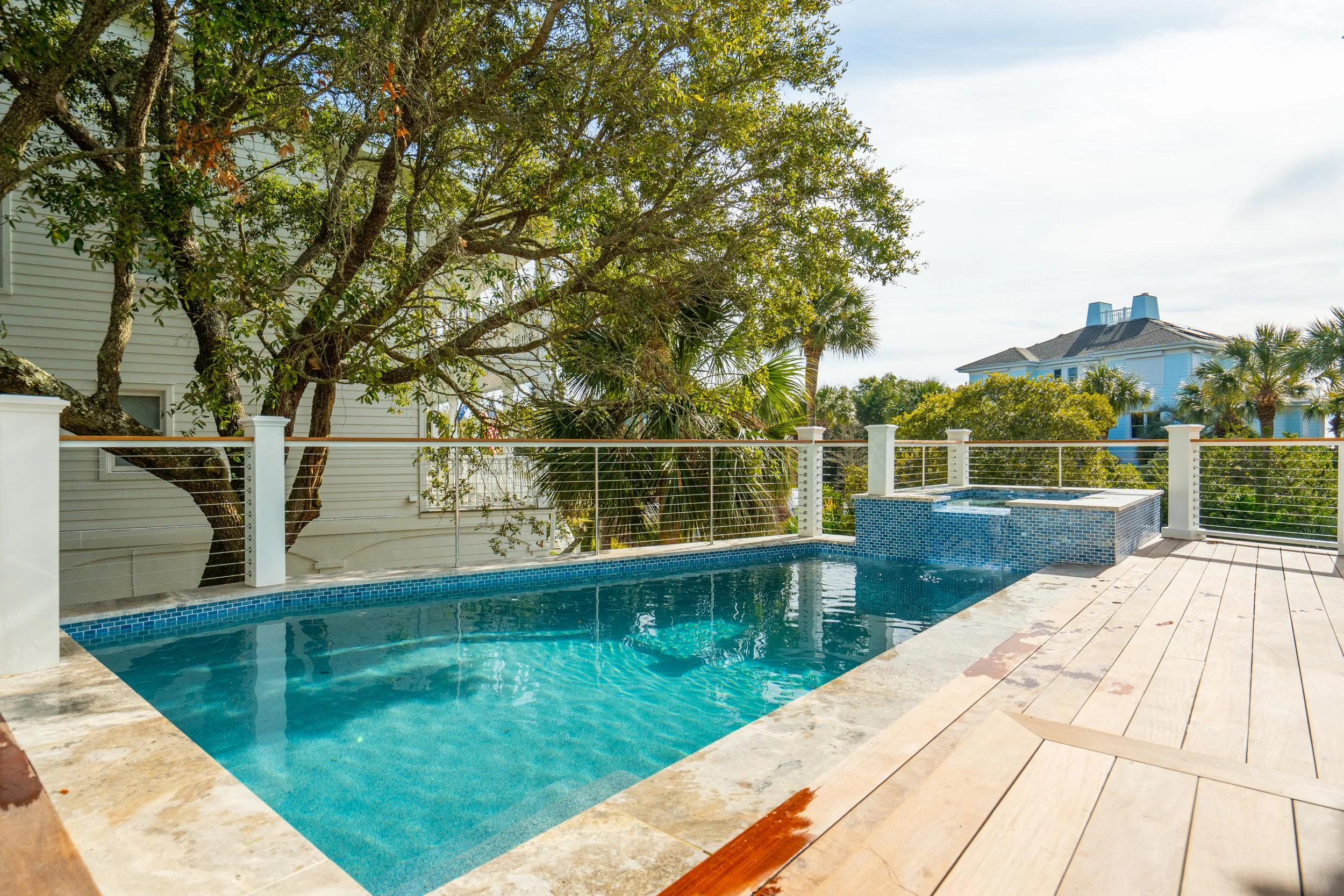 A backyard pool area with a wooden deck, a wire fence, and a hot tub, surrounded by lush trees and a neighboring house in the background.