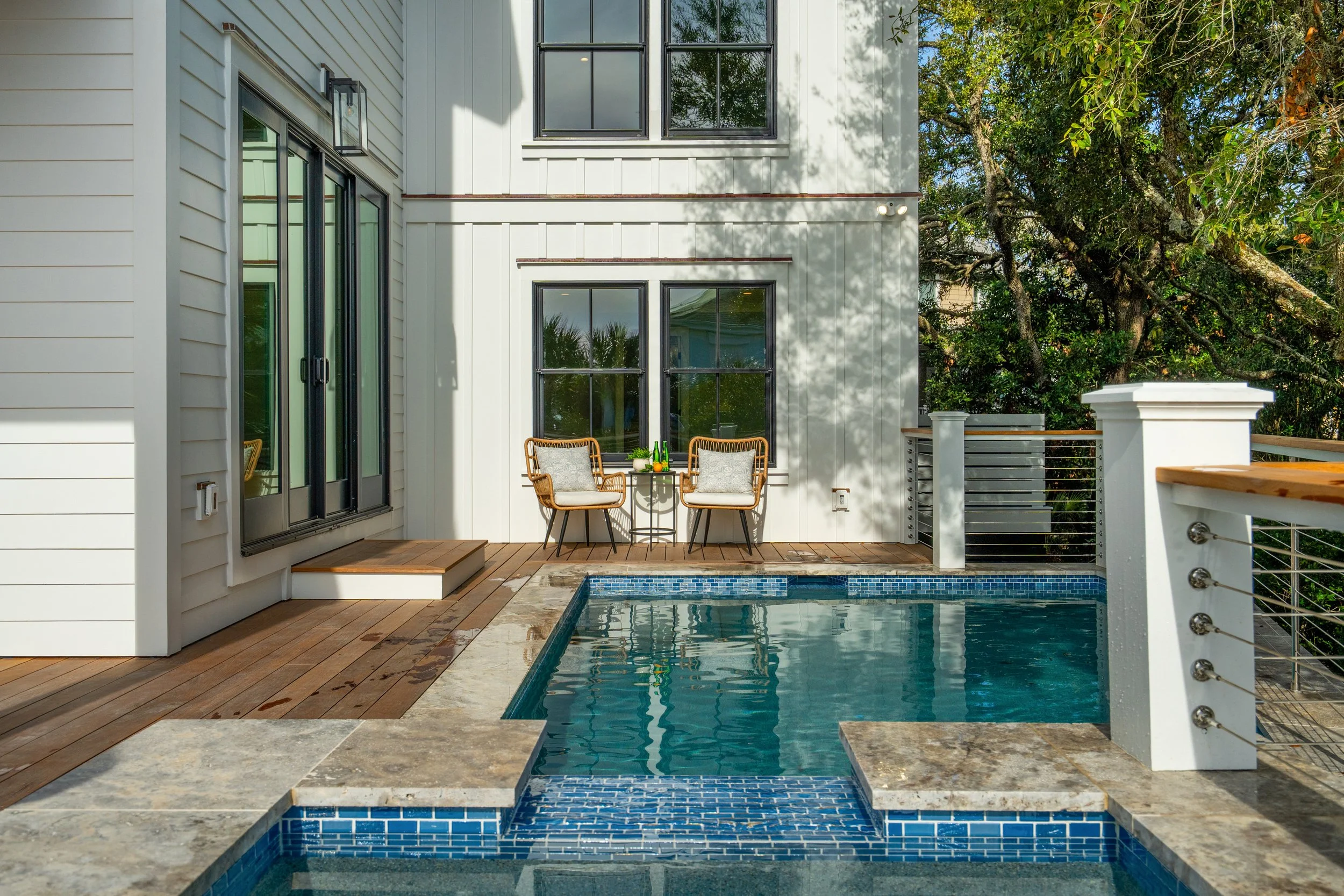 A backyard patio with a small pool, outdoor seating, and a white house with large windows surrounded by trees.