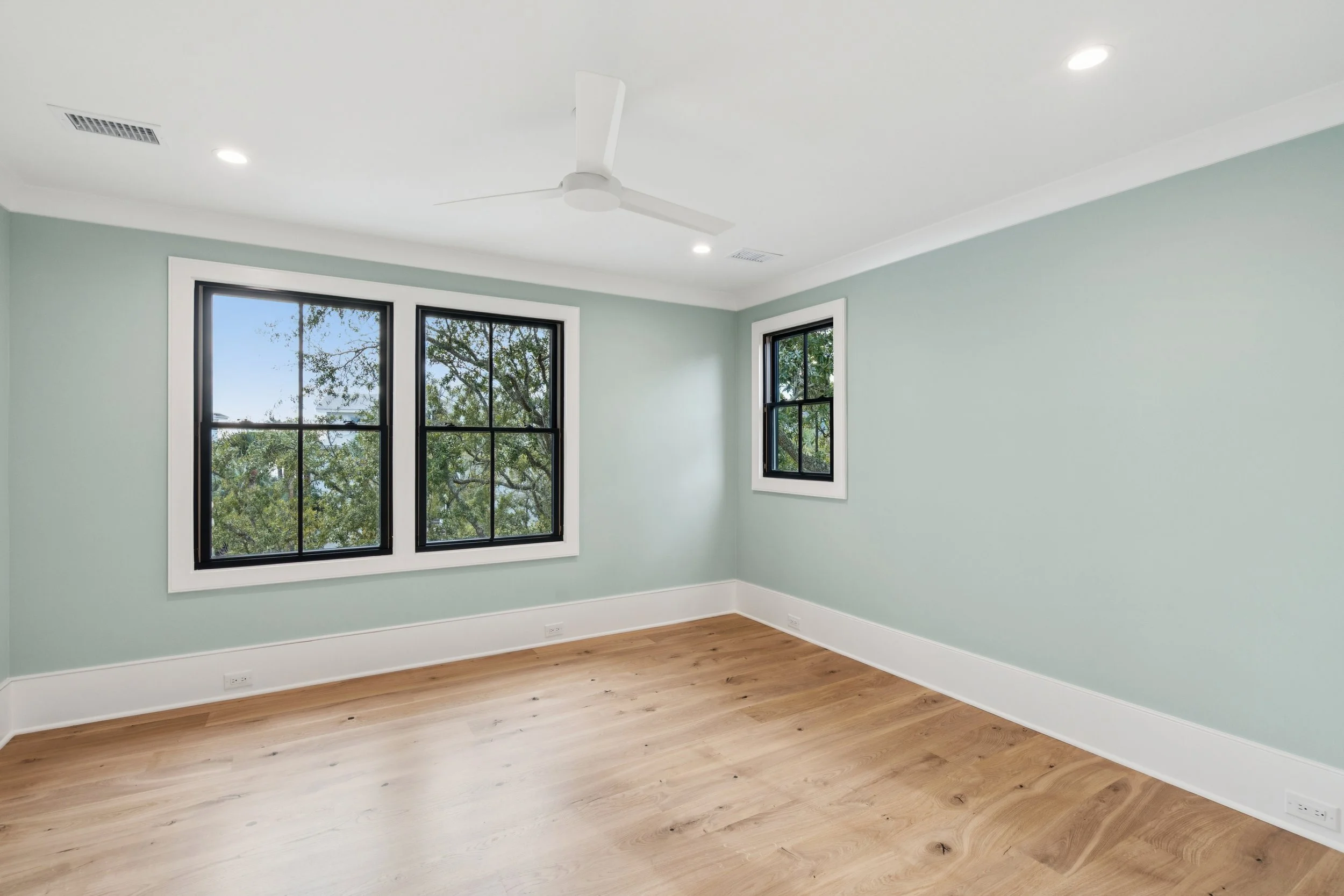 Empty room with hardwood floors, light green walls, three black-framed windows, white ceiling fan, recessed lighting, and white baseboards.