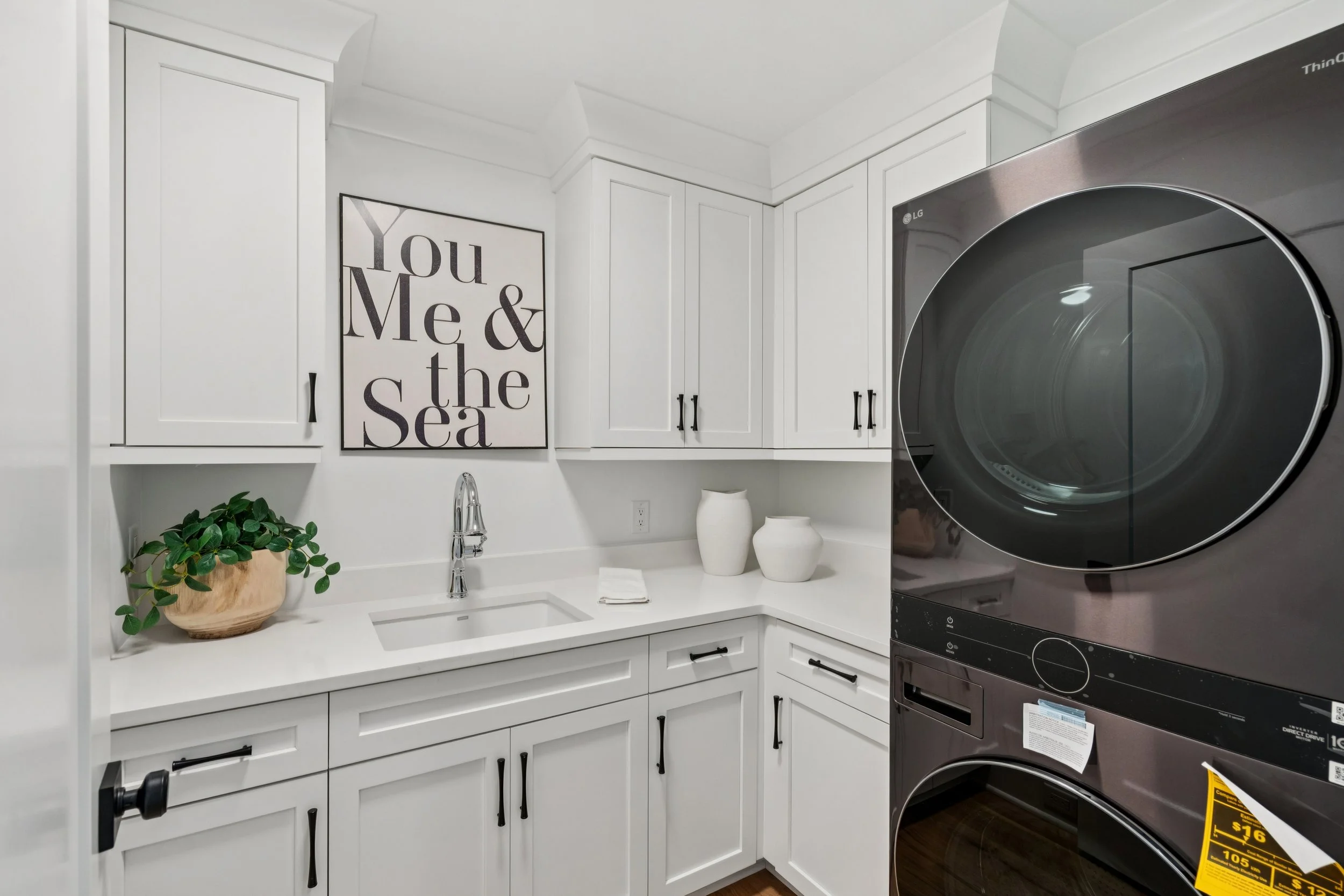 Laundry room with white cabinets, a small sink, a potted plant, and two white vases. A black and gray LG washer and dryer stack on the right. Wall art that reads 'You Me & the Sea' is on the wall above the sink.