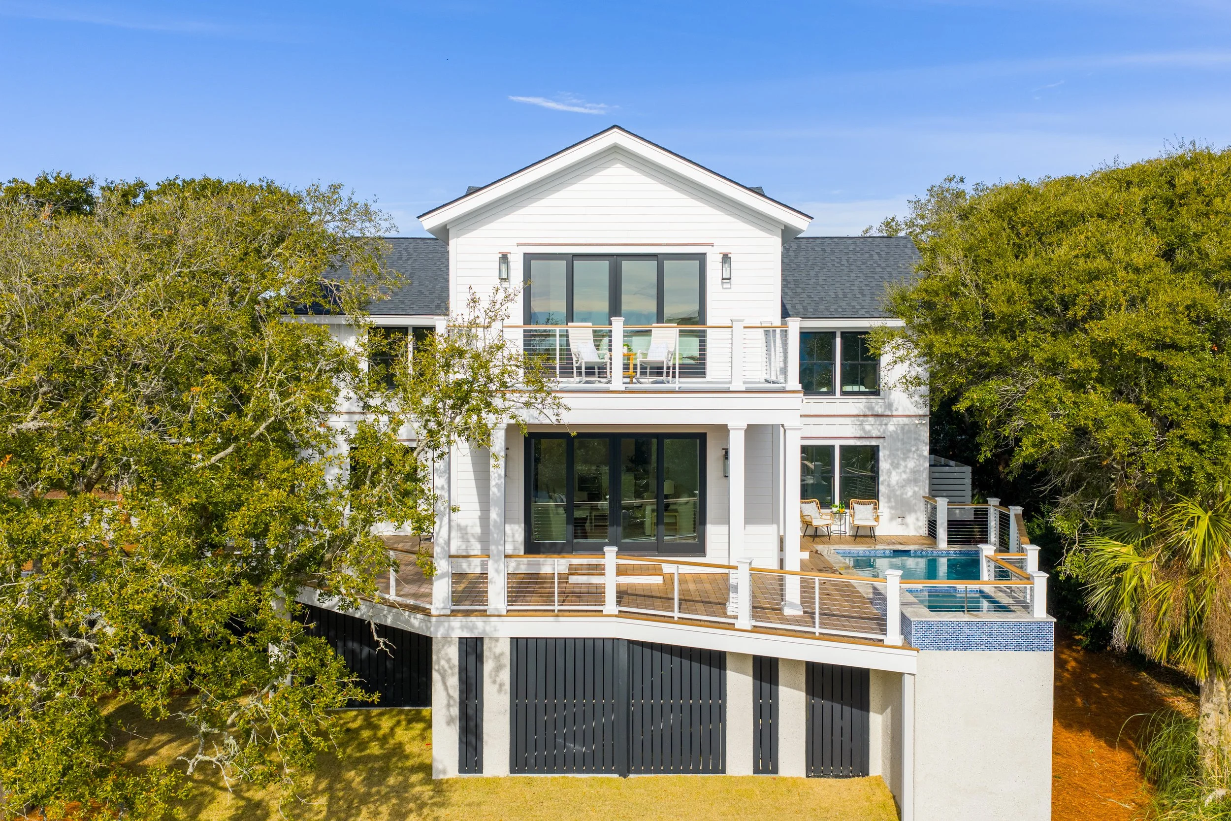 Modern two-story white house with large windows, surrounded by green trees, with decks and a swimming pool in backyard under a blue sky.