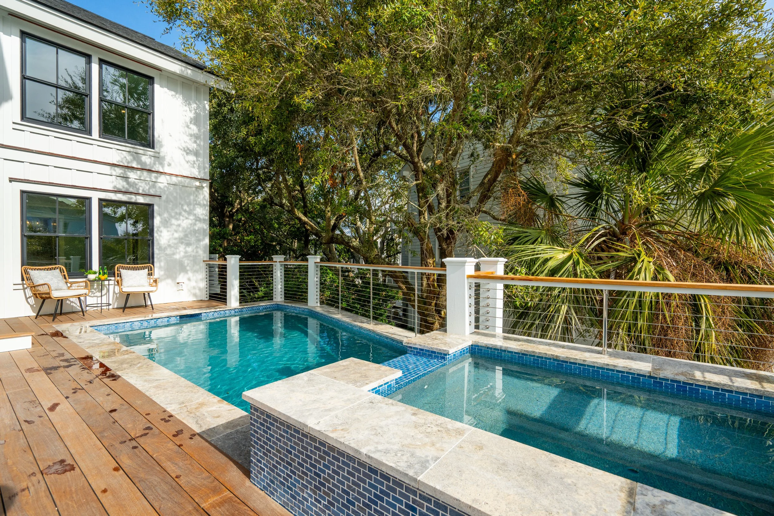 A backyard with a swimming pool next to a white house, surrounded by trees and foliage, with two chairs and a table on the wooden deck.