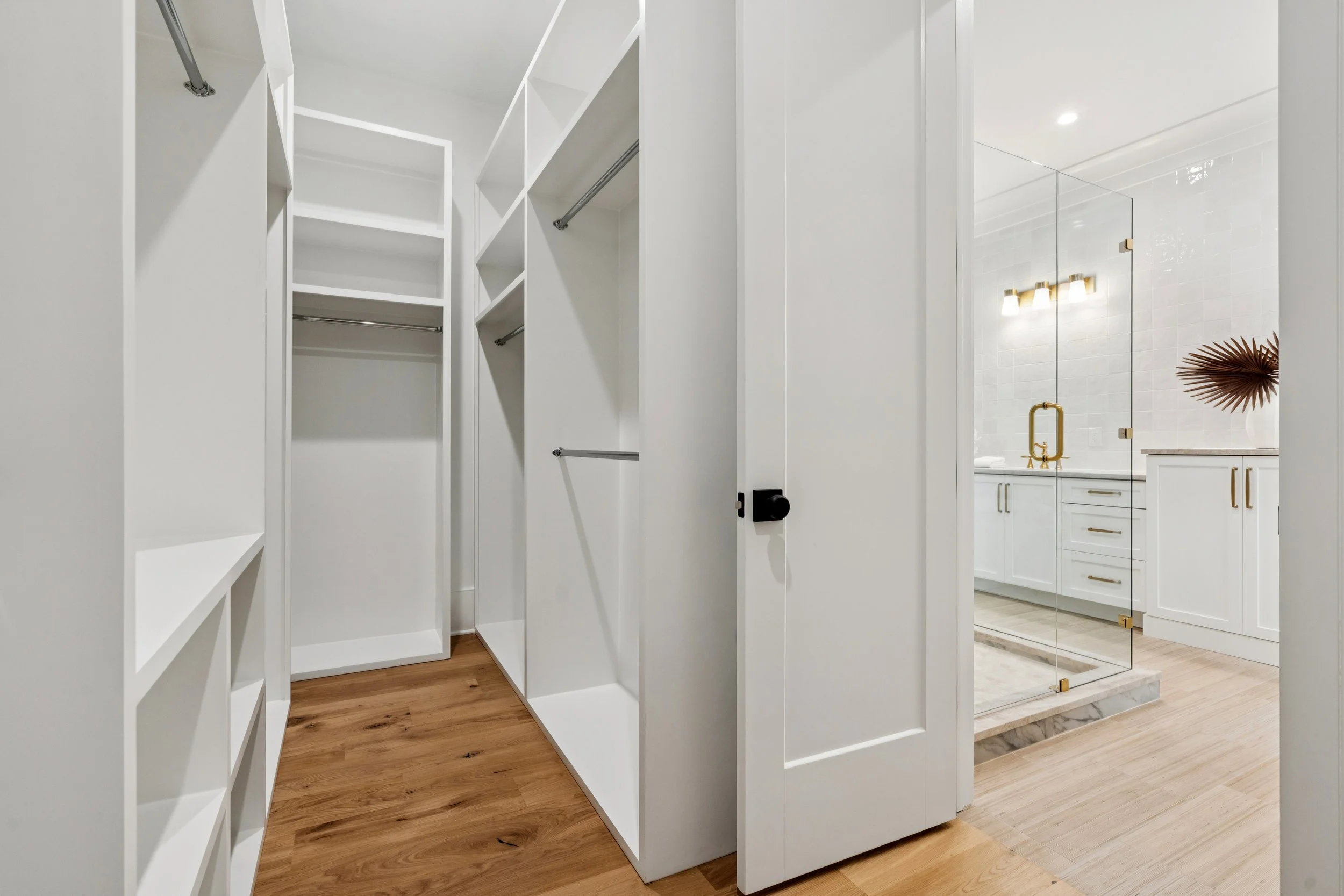 Empty walk-in closet with white built-in shelves and hanging rods, adjacent door leading to a modern bathroom with white cabinetry, gold fixtures, mirror, and decorative vase with dried palm leaves.