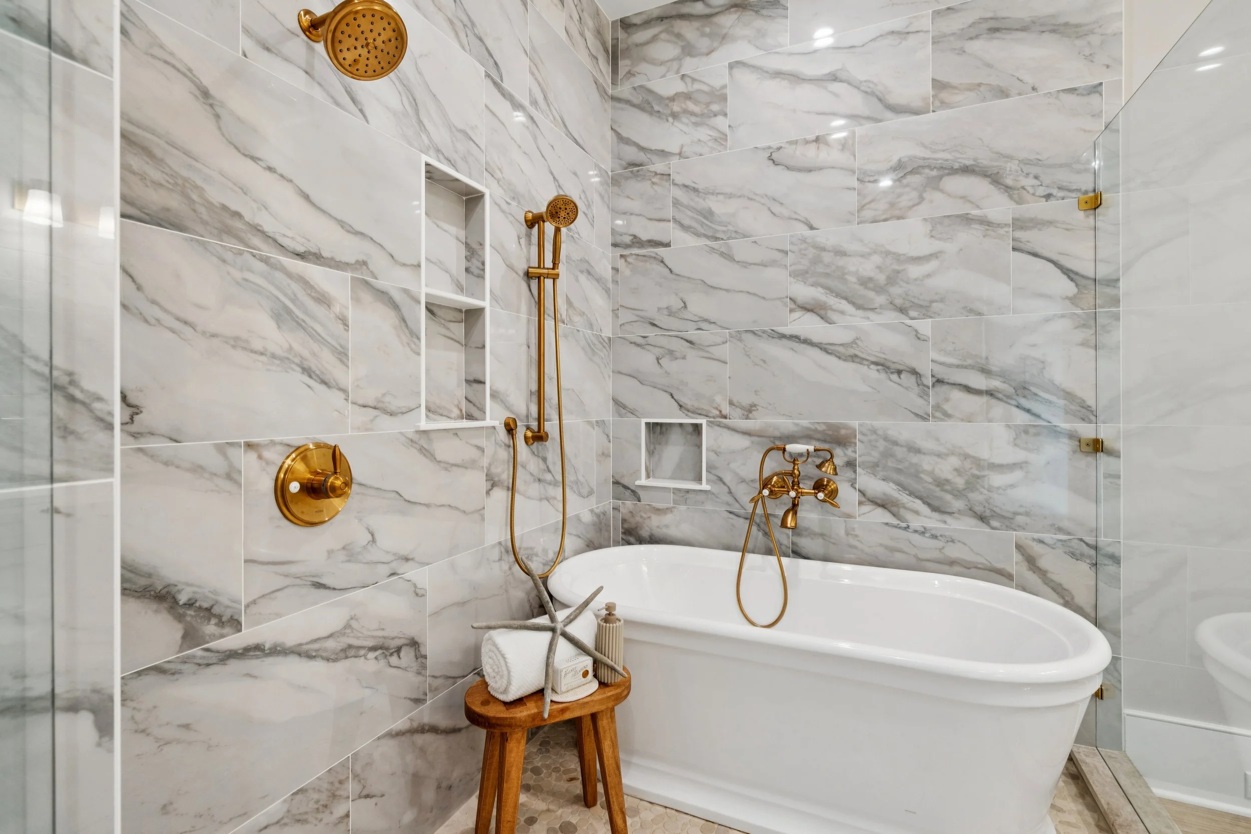 Elegant bathroom with marble walls, a white bathtub, gold fixtures, and a small wooden stool holding towels and decor.