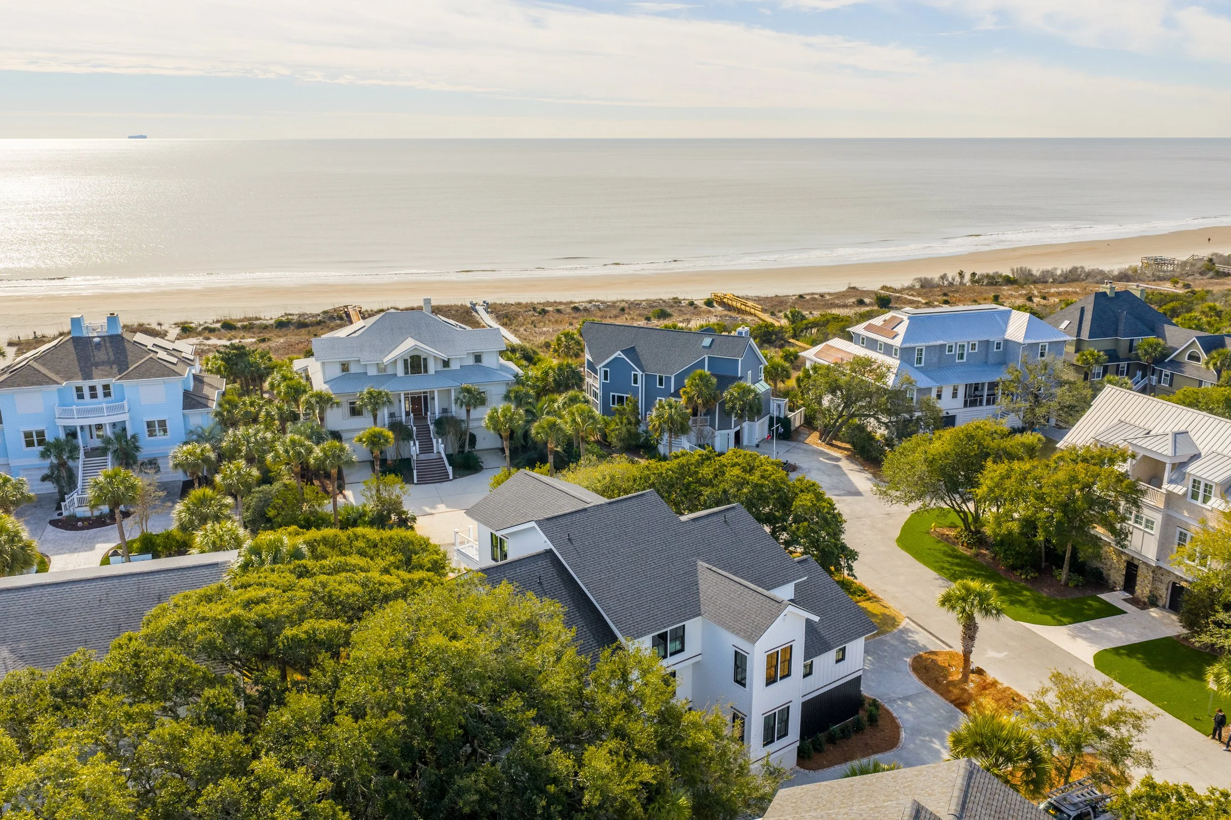 Aerial view of beachfront houses with trees, driveways, and a sandy beach along the ocean under a partly cloudy sky.