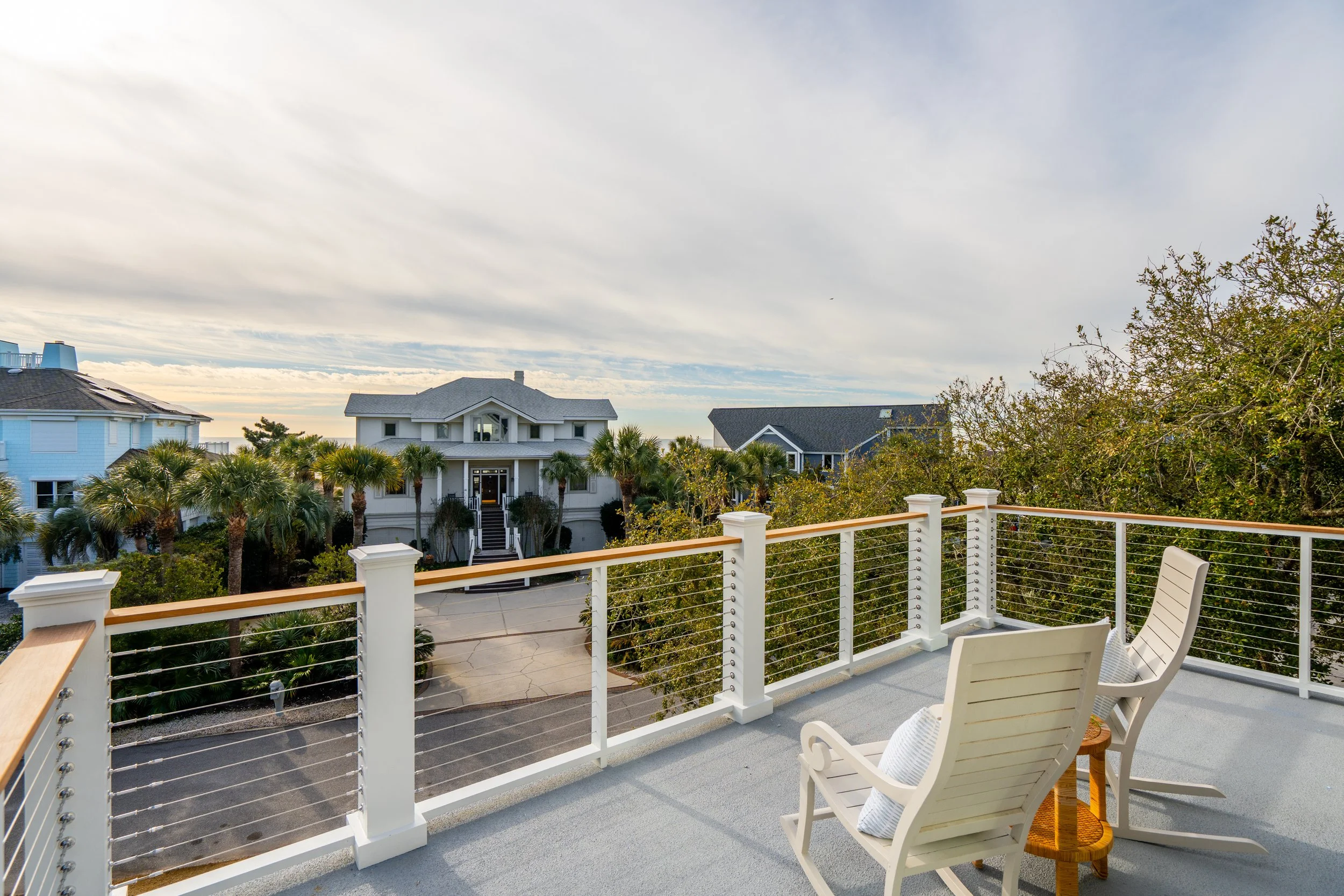 Balcony view with two white chairs and pillows overlooking palm trees and houses with gray roofs in a coastal neighborhood during cloudy daylight.