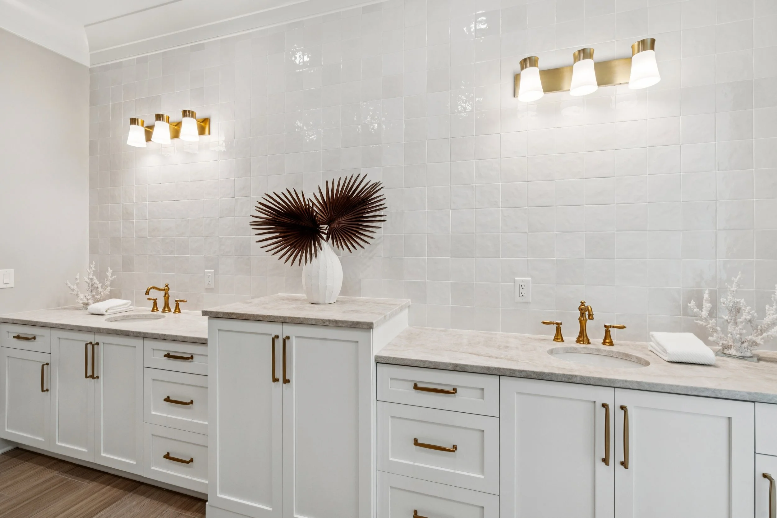 Bathroom with white cabinetry, marble countertops, gold fixtures, and a large white vase with dark palm leaves, illuminated by modern wall-mounted light fixtures.