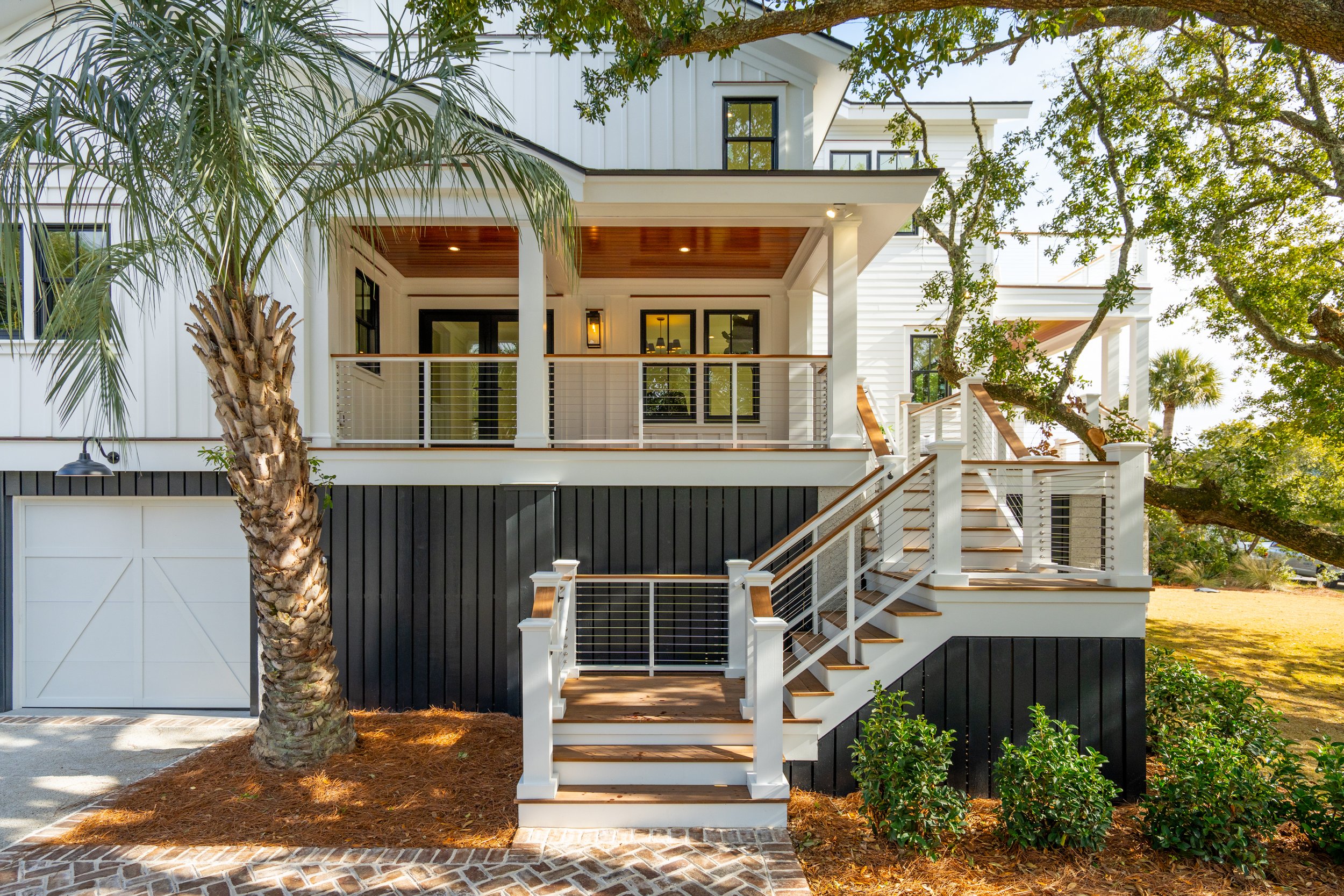 Front view of a modern multi-story house with a staircase leading to the front door, surrounded by greenery and trees, featuring white and dark exterior siding and a wooden porch ceiling.