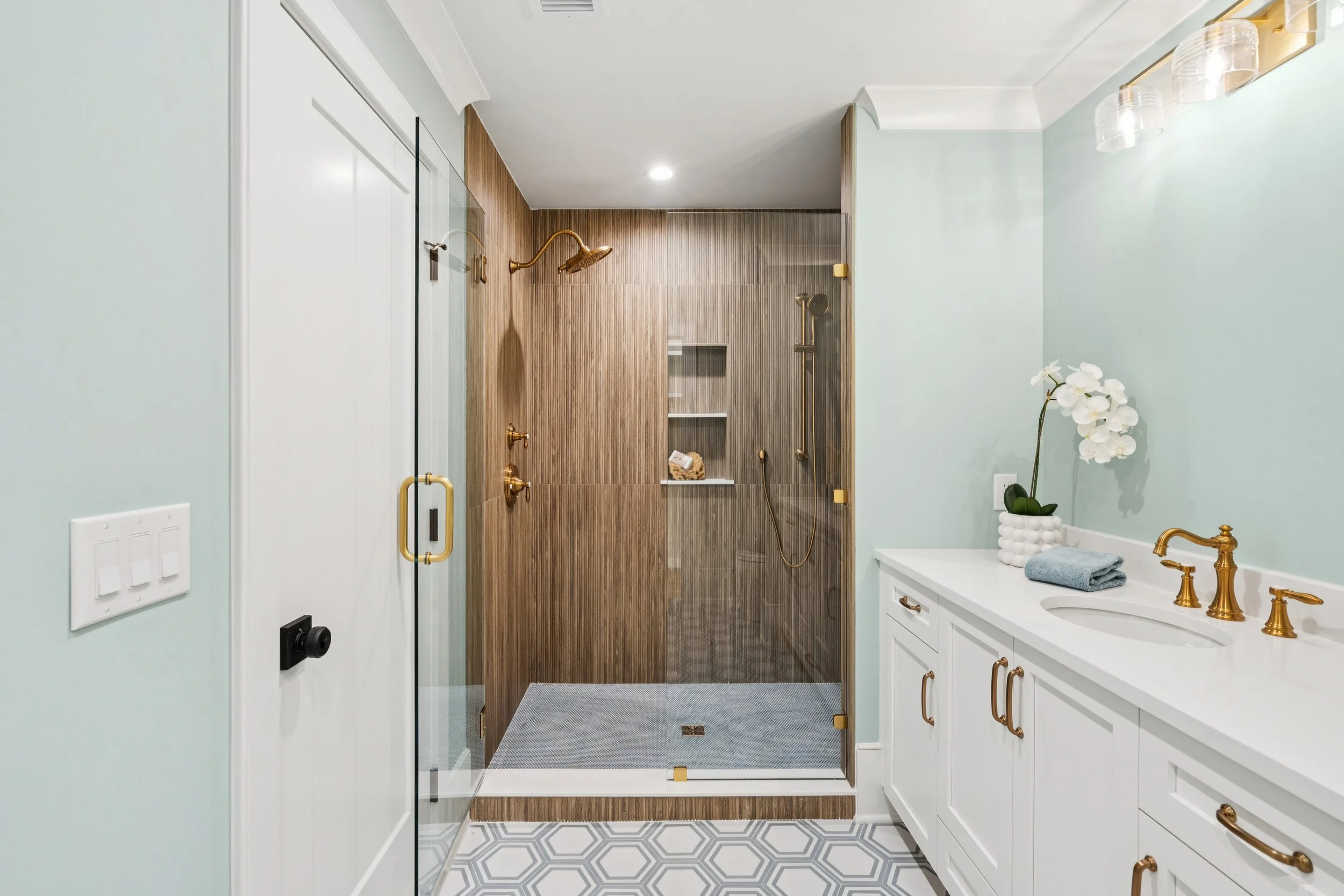 Modern bathroom with a glass-enclosed shower featuring wood-textured wall tiles, a white vanity with gold hardware, and a white orchid plant on the countertop.