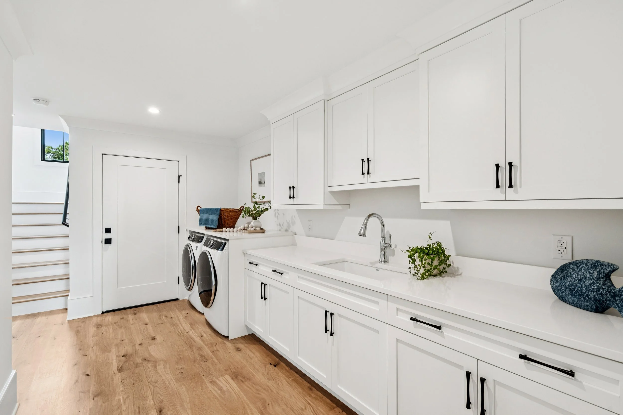 Laundry room with white cabinets, a sink, a washing machine and dryer, wooden flooring, decorative plants, and stairway in the background.