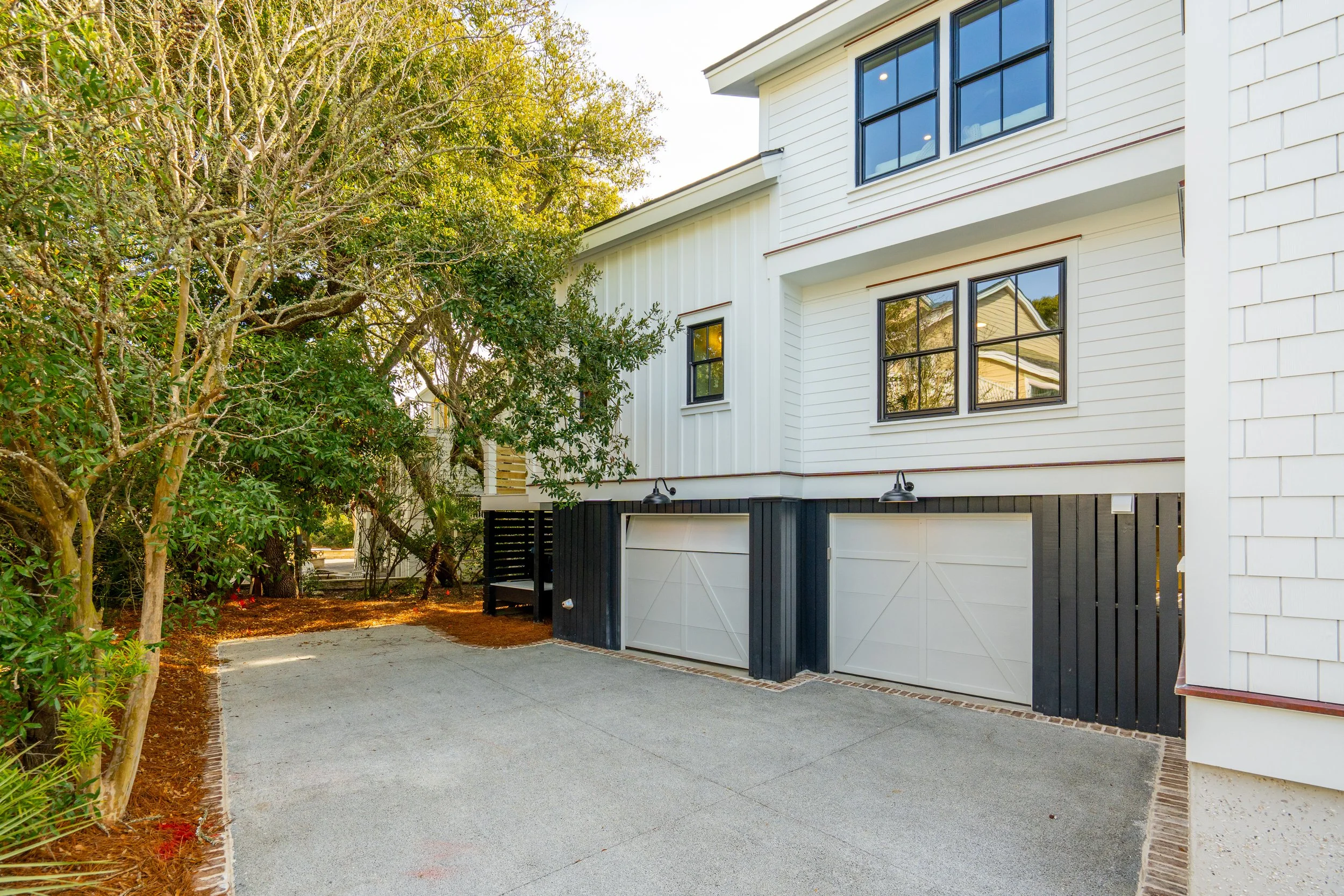 Exterior of a modern two-story house with a driveway, white siding, black window frames, and black and white garage doors, with trees nearby and outdoor lighting.