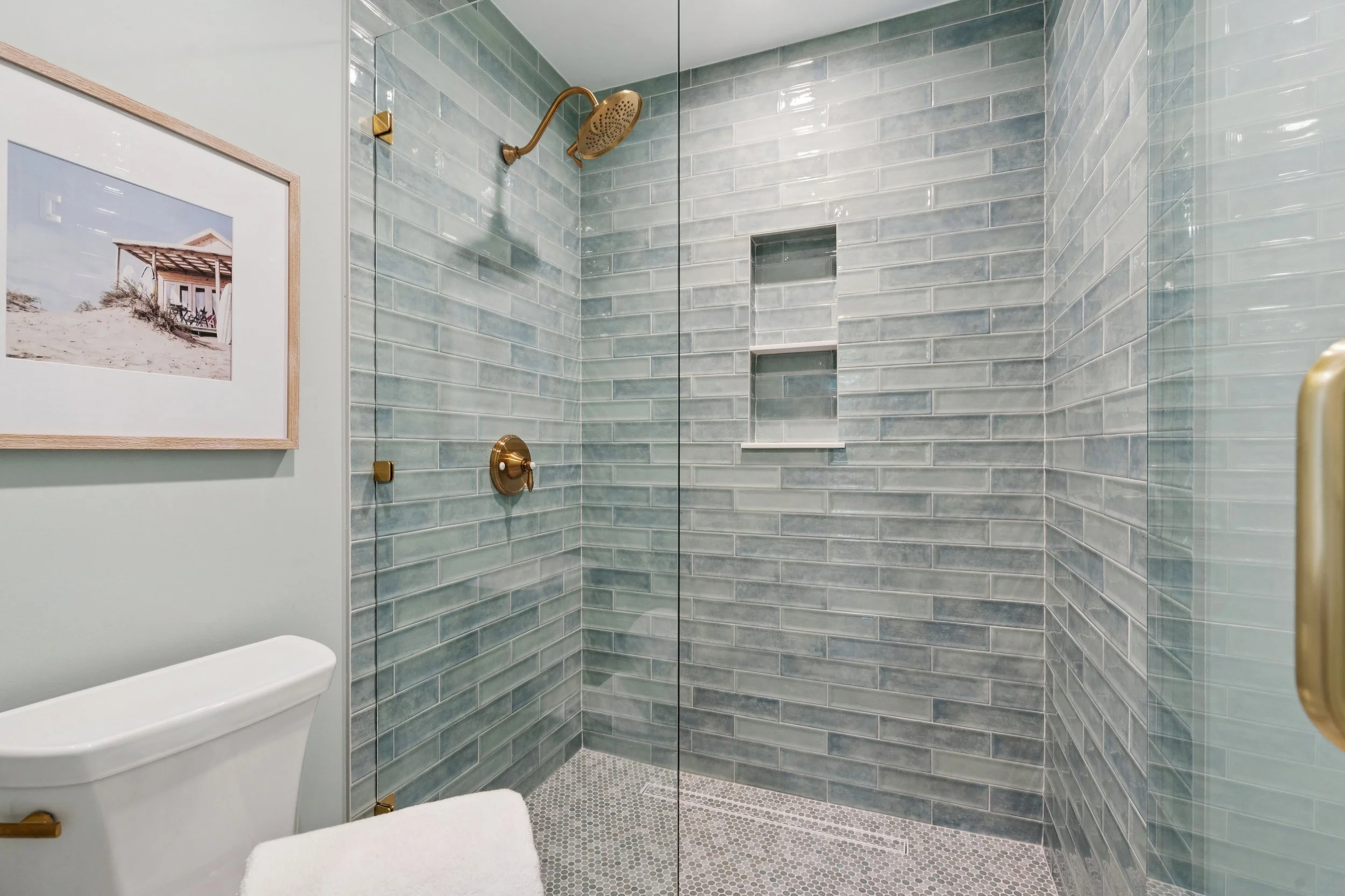 Modern bathroom with glass shower enclosure, grey subway tile walls, built-in shower shelves, and brass fixtures, including a shower head and handle, with a framed beach scene photo on the wall leaning against a white toilet.