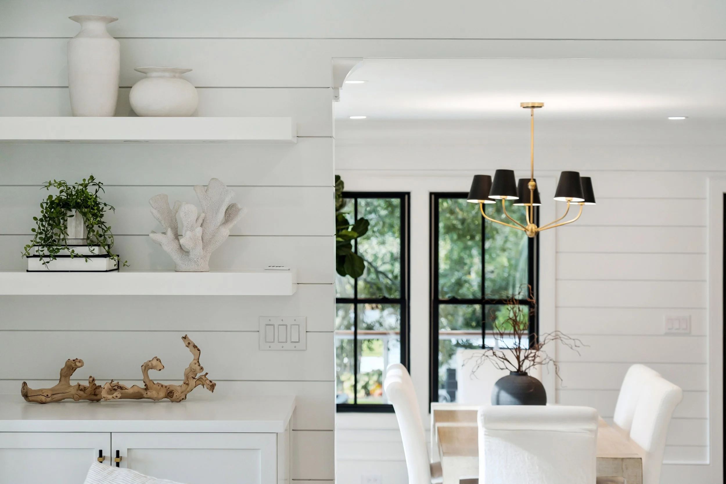 Modern living room and dining area with white shiplap walls, black-framed windows showing greenery outside, a gold chandelier with black shades over a dining table, decorative vases, coral sculpture, and driftwood art on shelves.