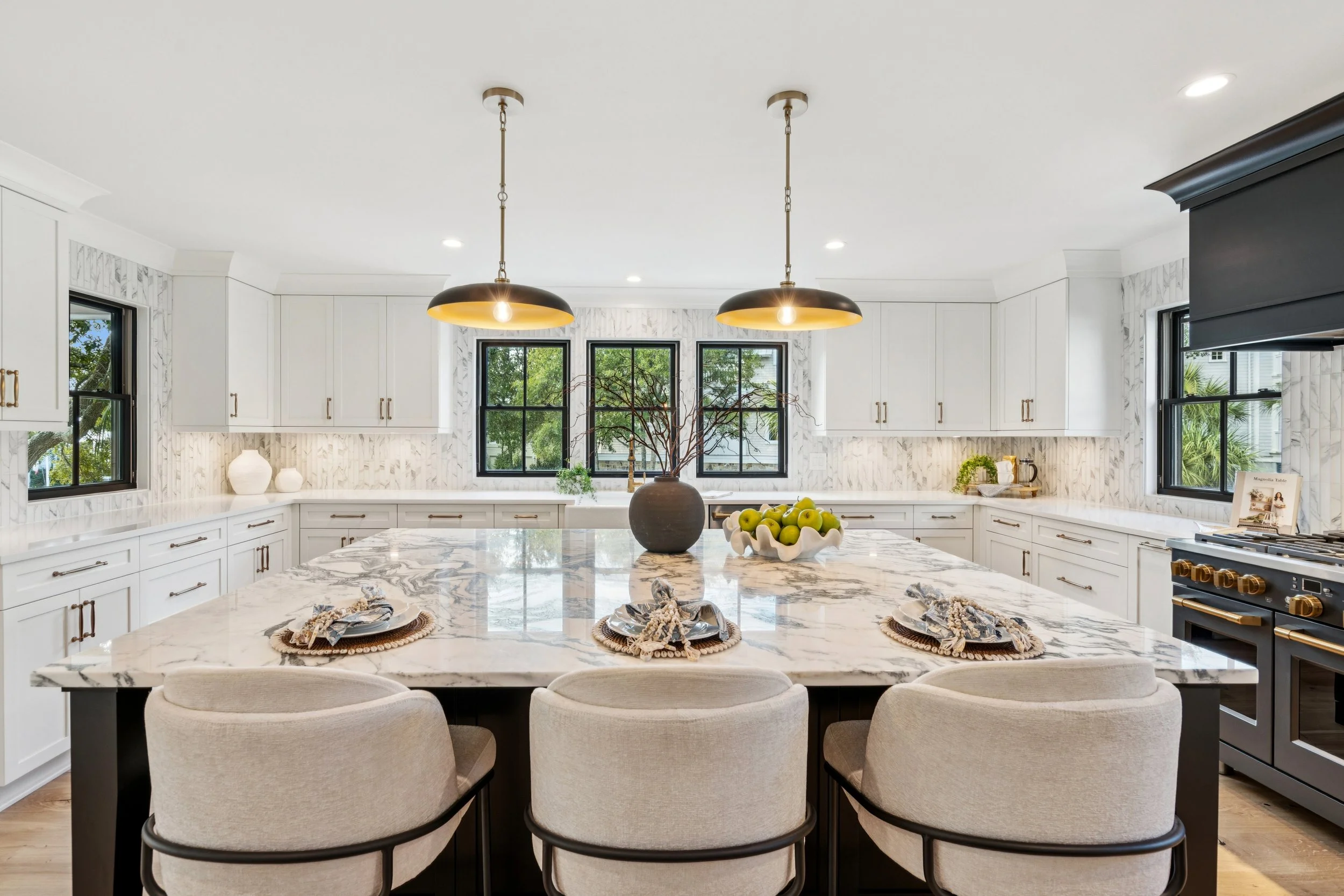Modern kitchen with white cabinets, marble countertops, skylight, black-framed windows, and an island with seating. Decor includes a black vase with branches and a white bowl of green apples.