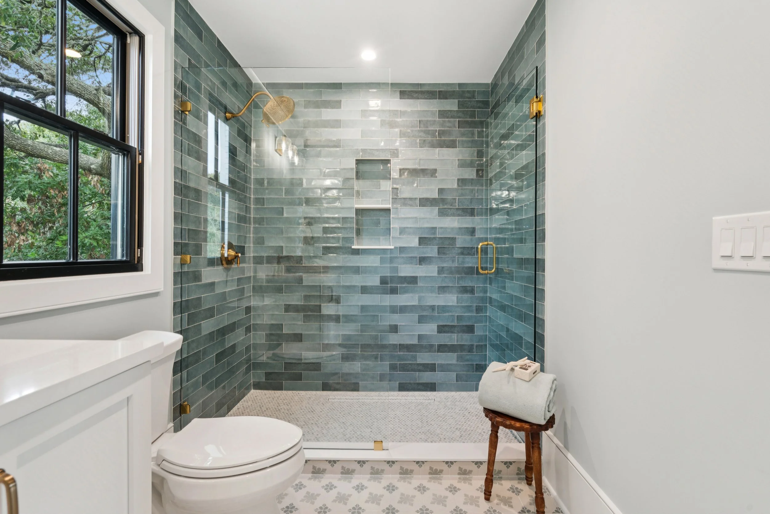 Bathroom with a walk-in shower featuring gray tile walls and gold fixtures, a window with a view of greenery, a white toilet, and a small stool with folded towels and a phone.