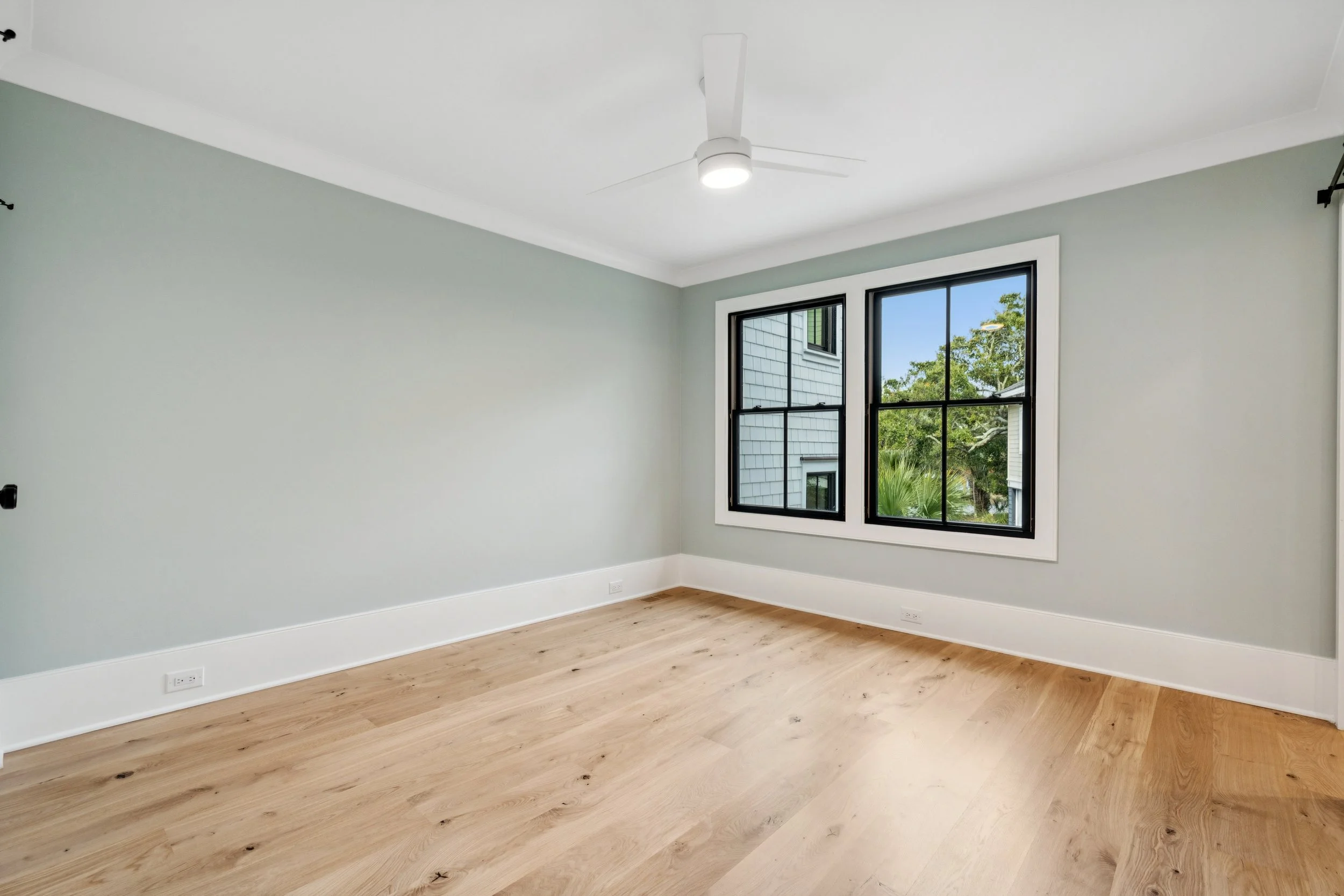 Empty room with light gray walls, hardwood flooring, a ceiling fan with light, and large black-framed windows showing green trees outside.