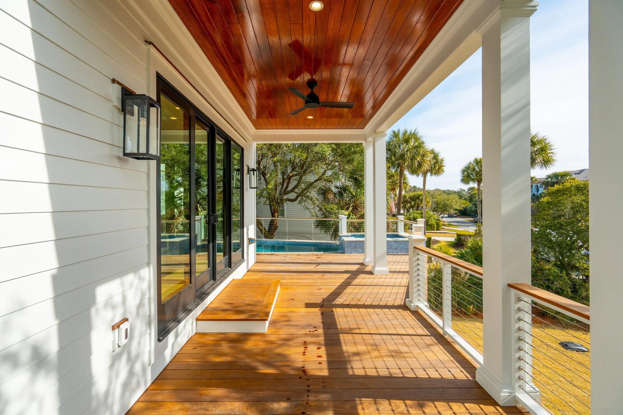 Covered balcony with wooden floor and ceiling, white columns, black ceiling fan, glass sliding doors, outdoor scenery with trees, palm trees, and neighboring houses, sunny weather.