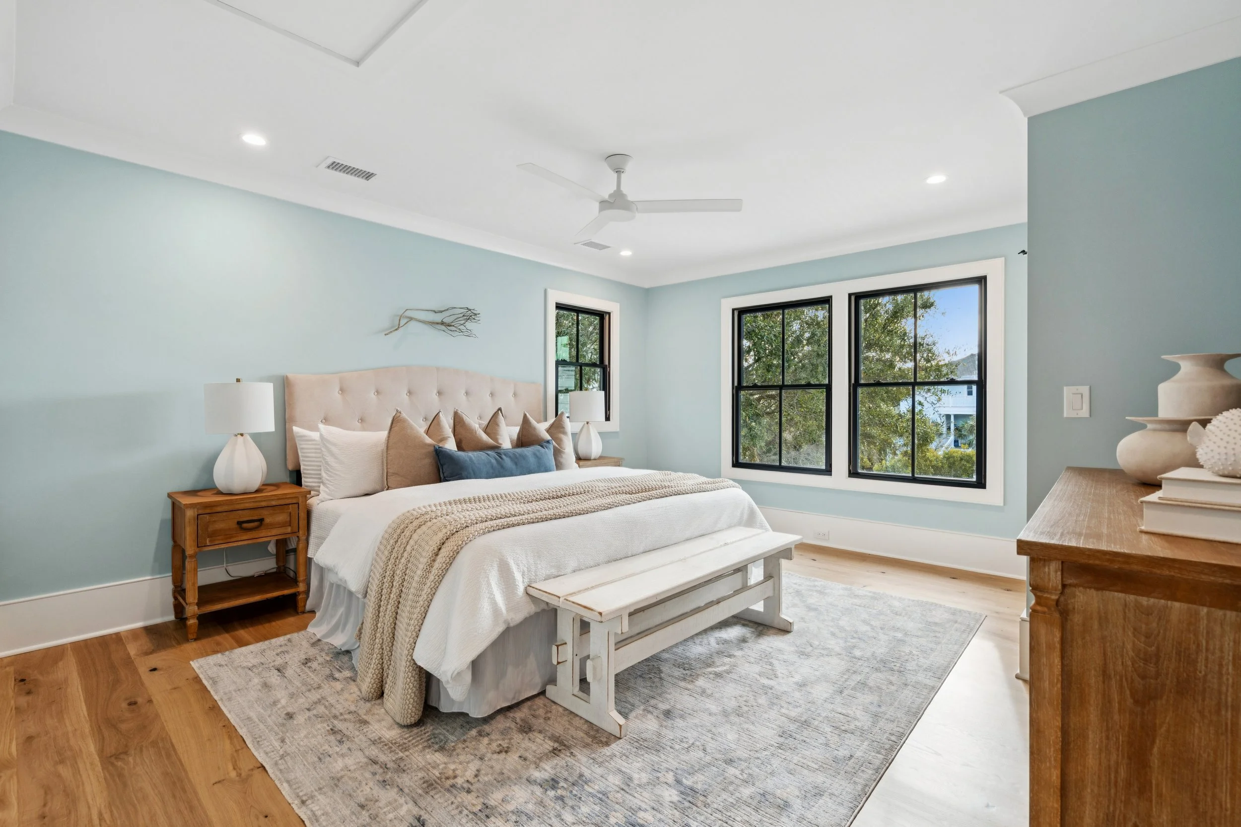 Bright bedroom with light blue walls, large black-framed windows, white bed with beige and blue pillows, wooden nightstands with white lamps, and a wooden dresser.