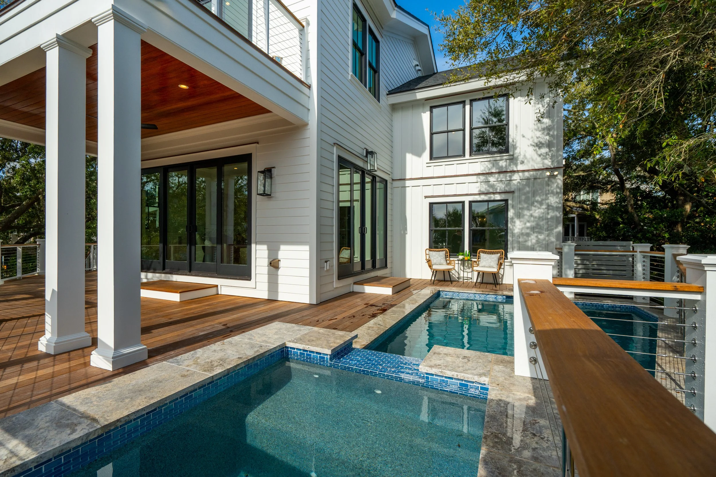 A modern house with a backyard pool area featuring wooden decking, white exterior walls with black French doors and large windows, a small seating area with two chairs, and shaded trees surrounding the space.