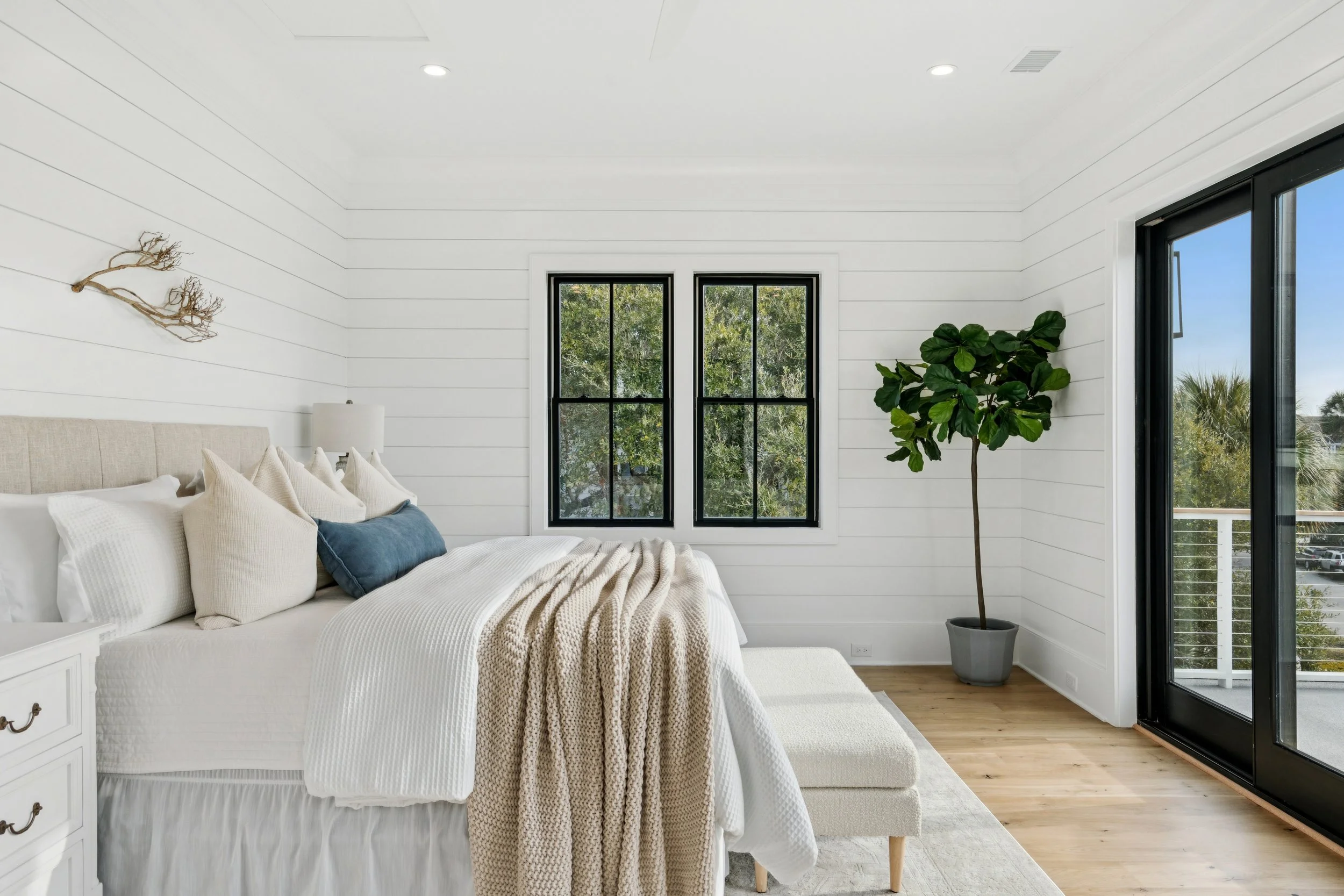 A bedroom with white walls, a bed with white and beige bedding, a beige headboard, a nightstand, a white lamp, a small bench, tiled flooring, two black-framed windows, a large potted plant, and a glass door leading to an outdoor balcony.