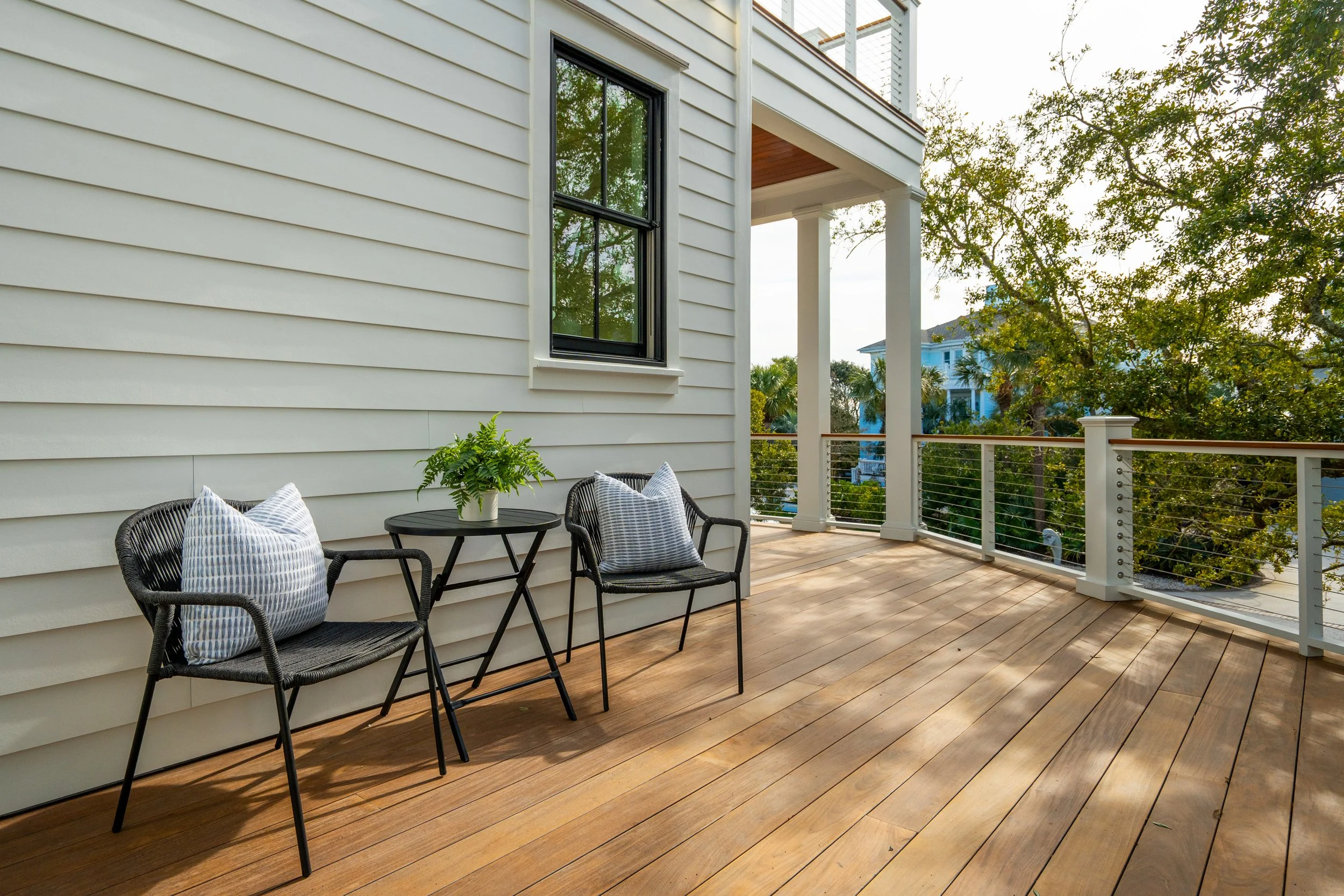 A spacious balcony with wooden flooring, two black chairs with pillows, a small table with a plant, surrounded by a white railing, with trees and houses visible in the background.