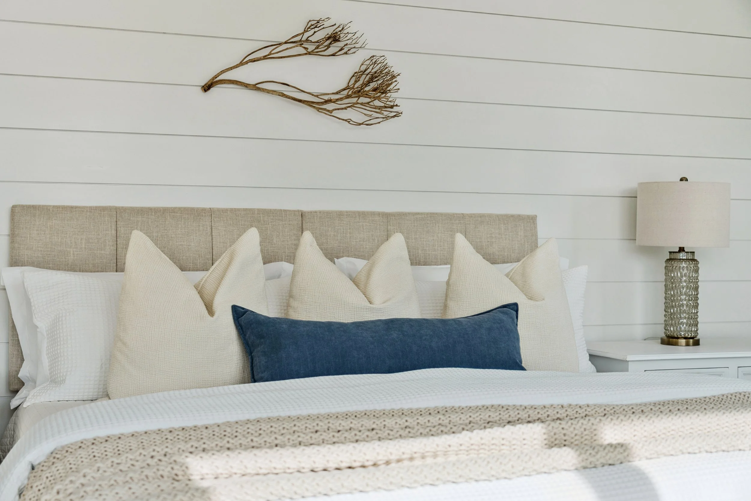 A bedroom with a beige upholstered headboard, multiple cream and beige pillows, a blue throw pillow, a white textured blanket, a white side table, and a lamp with a textured glass base and a beige lampshade. A dried branch art piece hangs above the b